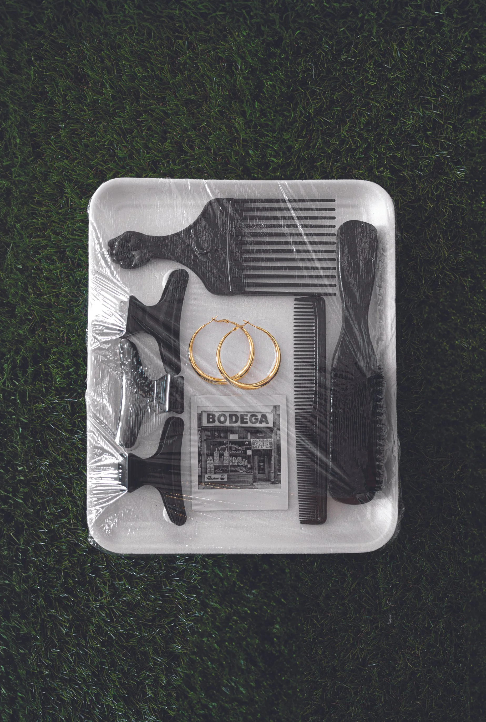 A package wrapped in clear plastic containing black hair-cutting tools, a pair of gold hoop earrings, a small magazine, a comb, and a hairbrush, all placed on a grassy surface.