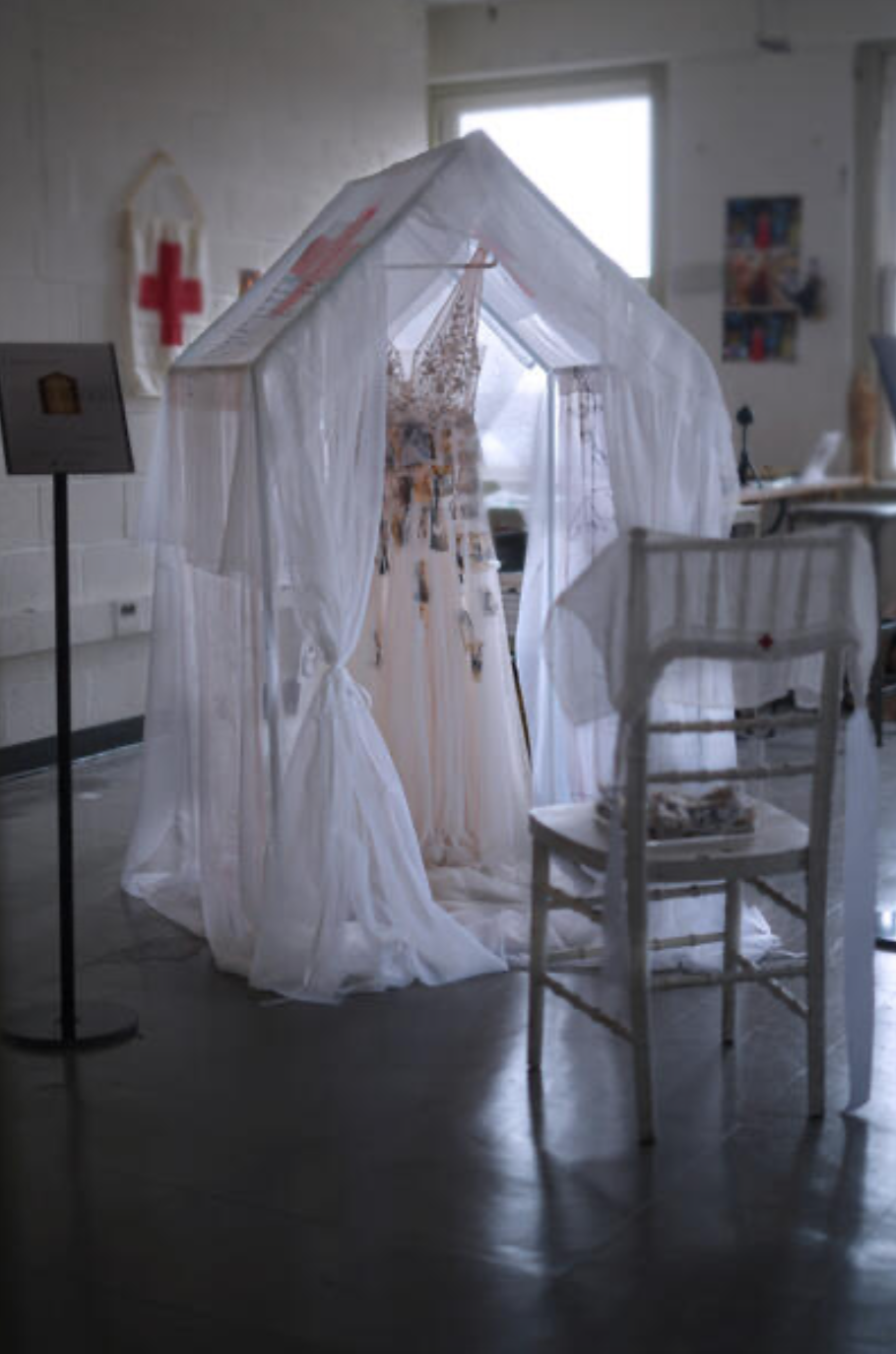 A white wedding dress on a mannequin inside a small white wedding chapel with sheer white curtains, surrounded by wedding-related decorations.
