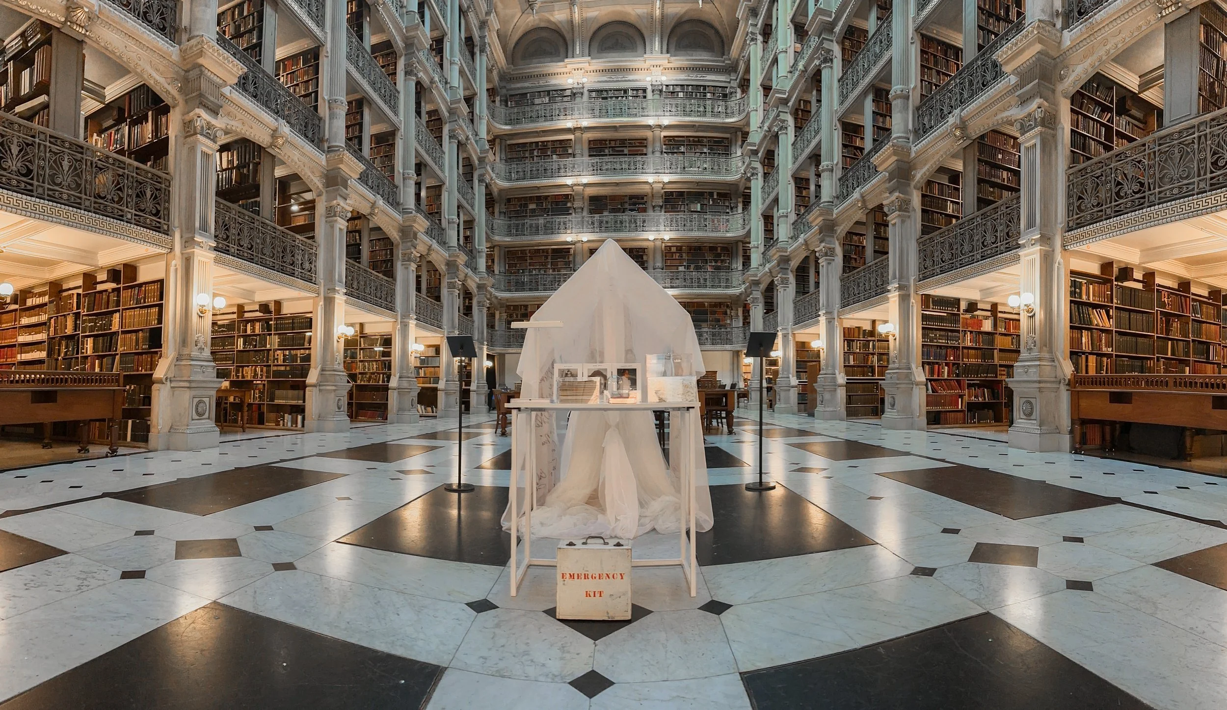 Inside a grand, historic library with multiple levels of bookshelves, ornate railings, and marble floors. In the center, there is a makeshift structure covered with a white cloth, with an emergency kit box in front of it.