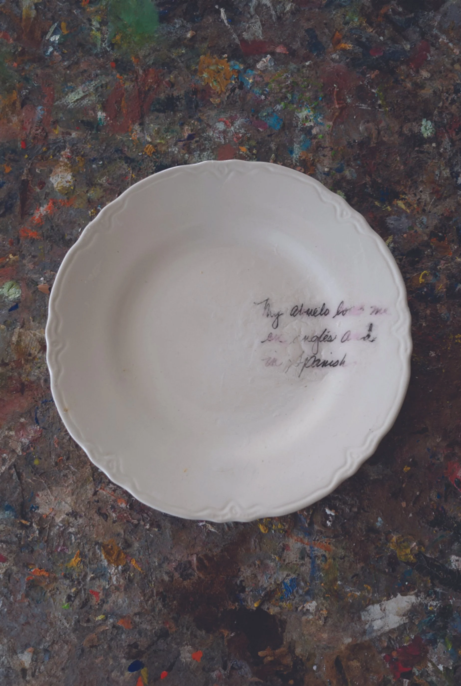 A white porcelain plate with a decorative scalloped edge, placed on a colorful, paint-splattered surface. There is handwritten text on the inside of the plate that reads, 'My alquido for me is en inglés a la español'.
