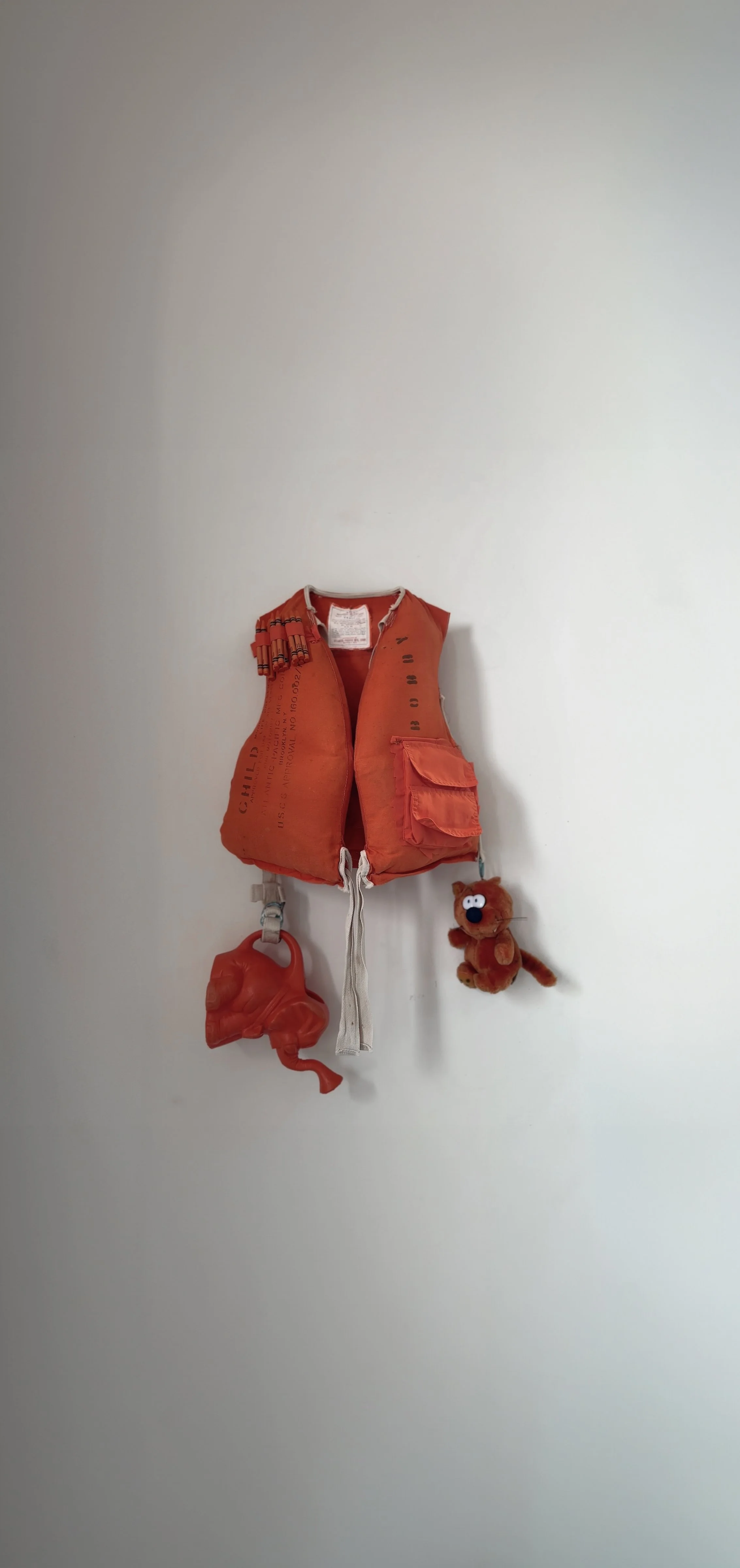 Child's orange life jacket hanging on a hook on a white wall, with a plush toy of a brown bear with googly eyes and a red tail, and a red plastic animal-shaped object hanging below.
