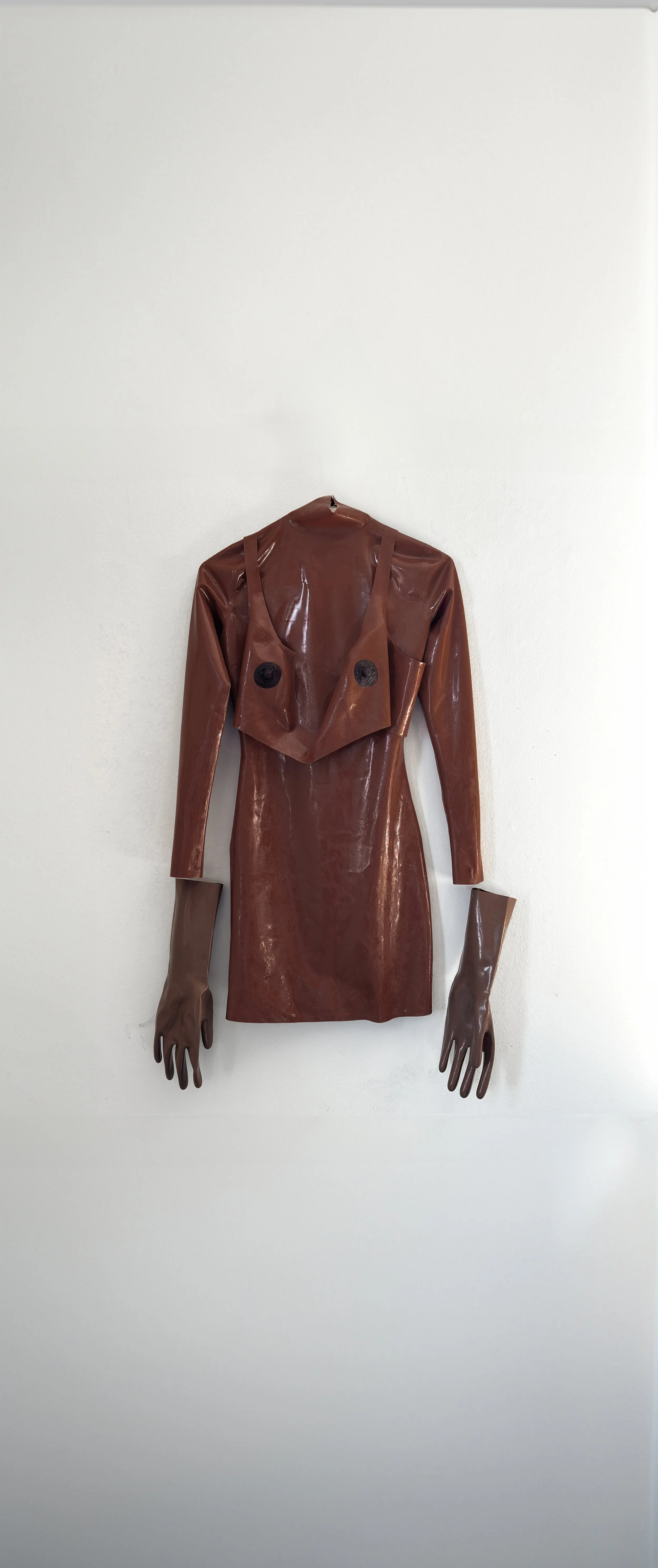 An art installation of a dress made from brown shiny material resembling leather, with matching gloves attached. The dress has an unusual design with a draped top and no head, face, or other visible body parts.