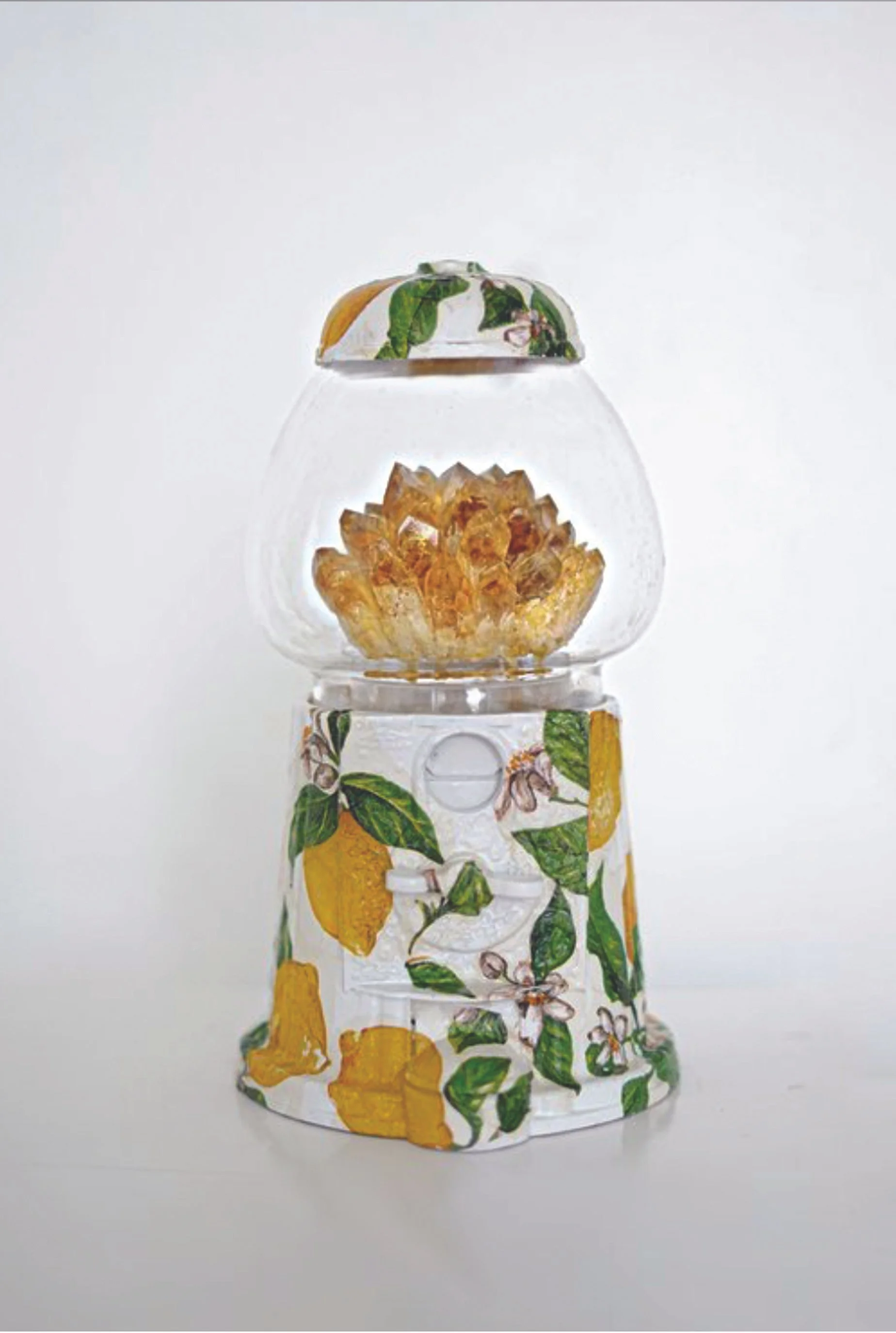 A glass citrus juicer with a lemon and lemon flower design, containing a sliced lemon in the top section and lemon zest in the middle section, against a plain background.