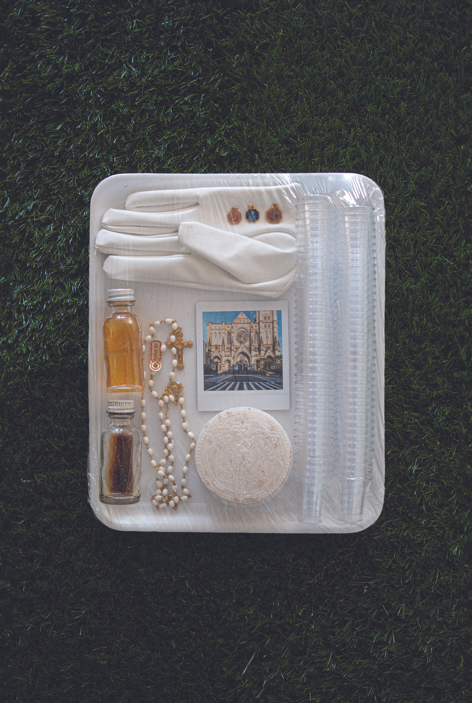 A tray with religious items including rosary beads, small bottles with liquids, a photograph of a church, a round white object, a glove, and small jewelry pieces, on a grassy surface.