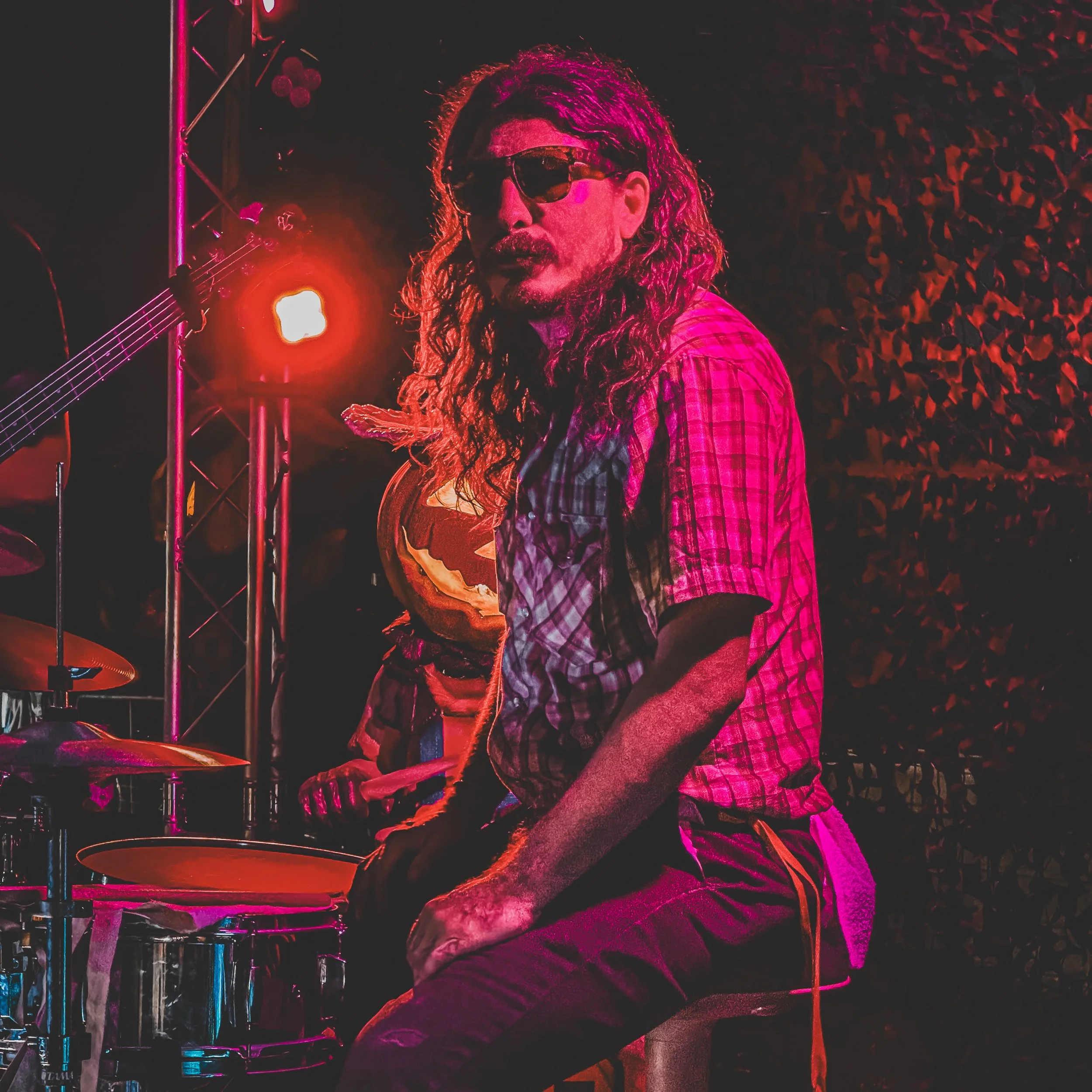A man with long curly hair, wearing sunglasses and a plaid shirt, sitting on a stool with a drum set in front of him. Bright red stage lighting is visible behind him.