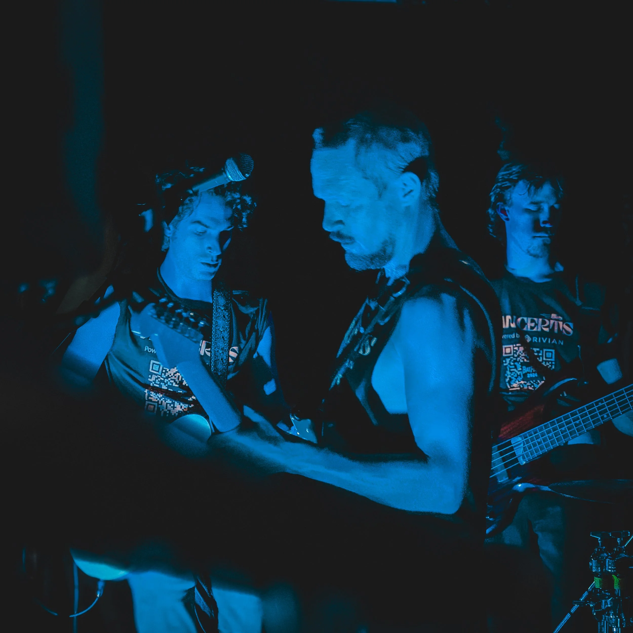 Three musicians on stage illuminated by blue light, one playing guitar and two with microphones, in a dark performance setting.