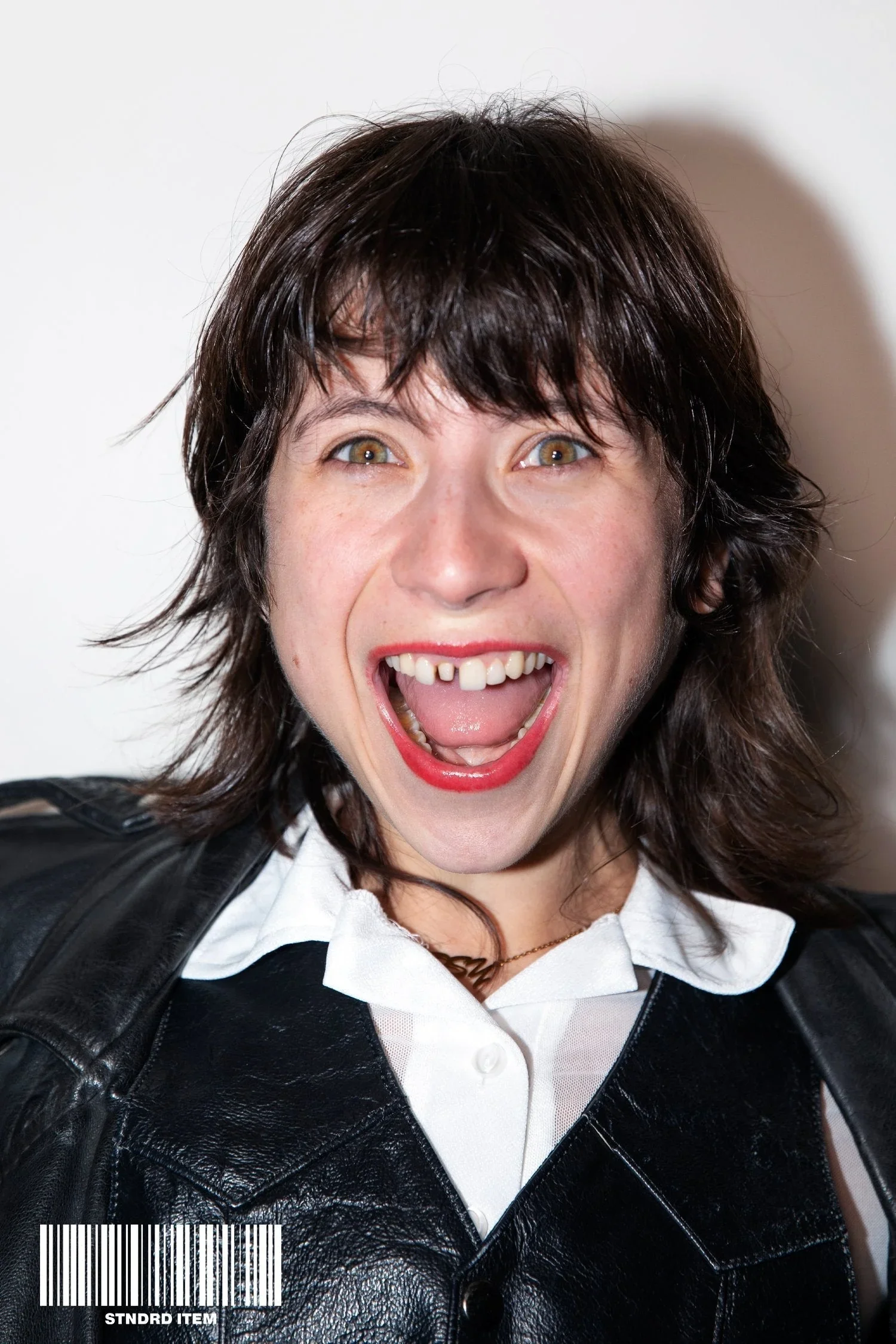A woman with dark brown, wavy hair smiling widely with her mouth open, showing her teeth. She's wearing a black leather jacket over a white shirt and red lipstick.