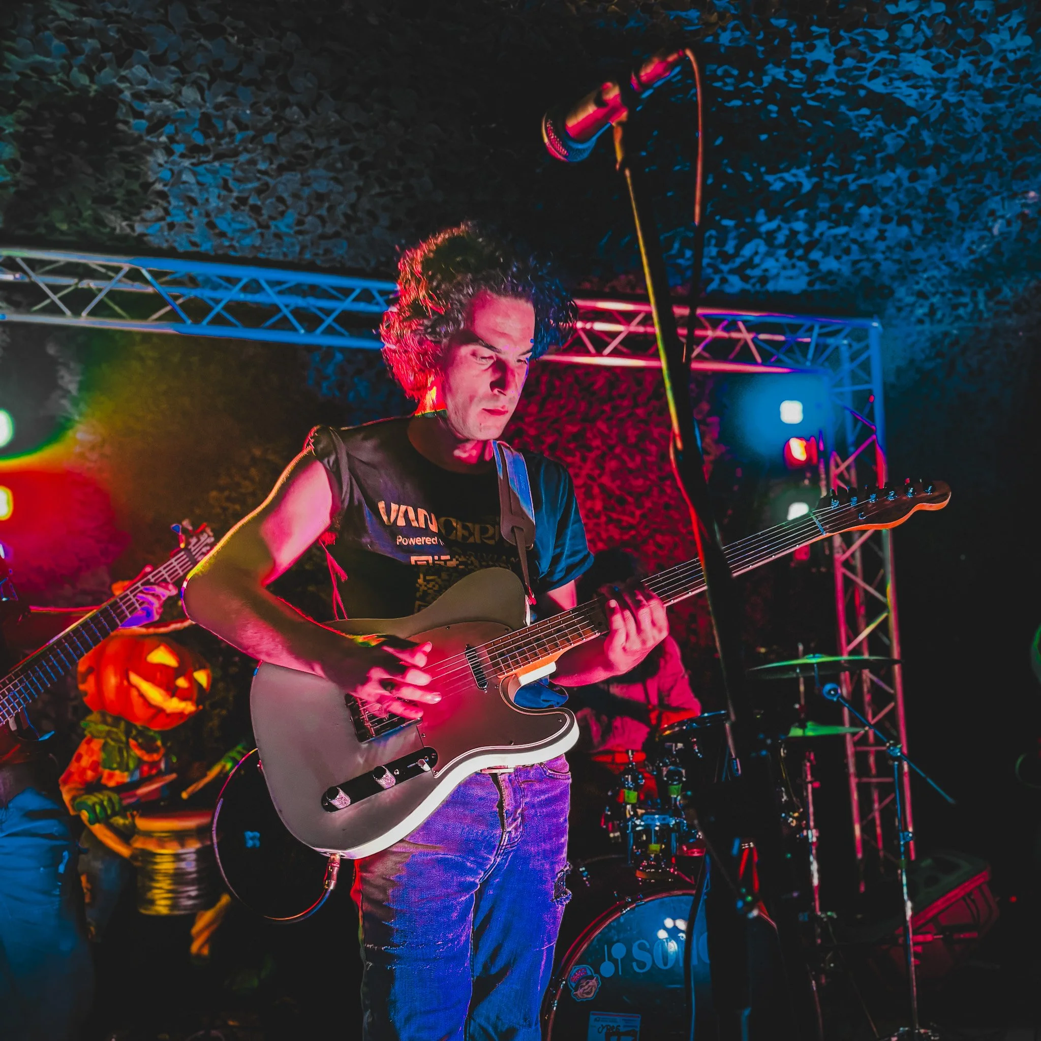A person playing an electric guitar on a stage at night, with colorful lights and a Halloween-themed decoration in the background.
