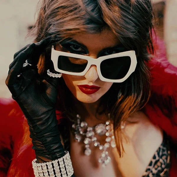 A woman with dark hair and red lipstick wearing large white sunglasses, black gloves, a diamond bracelet, and jewelry, posing with a confident expression.