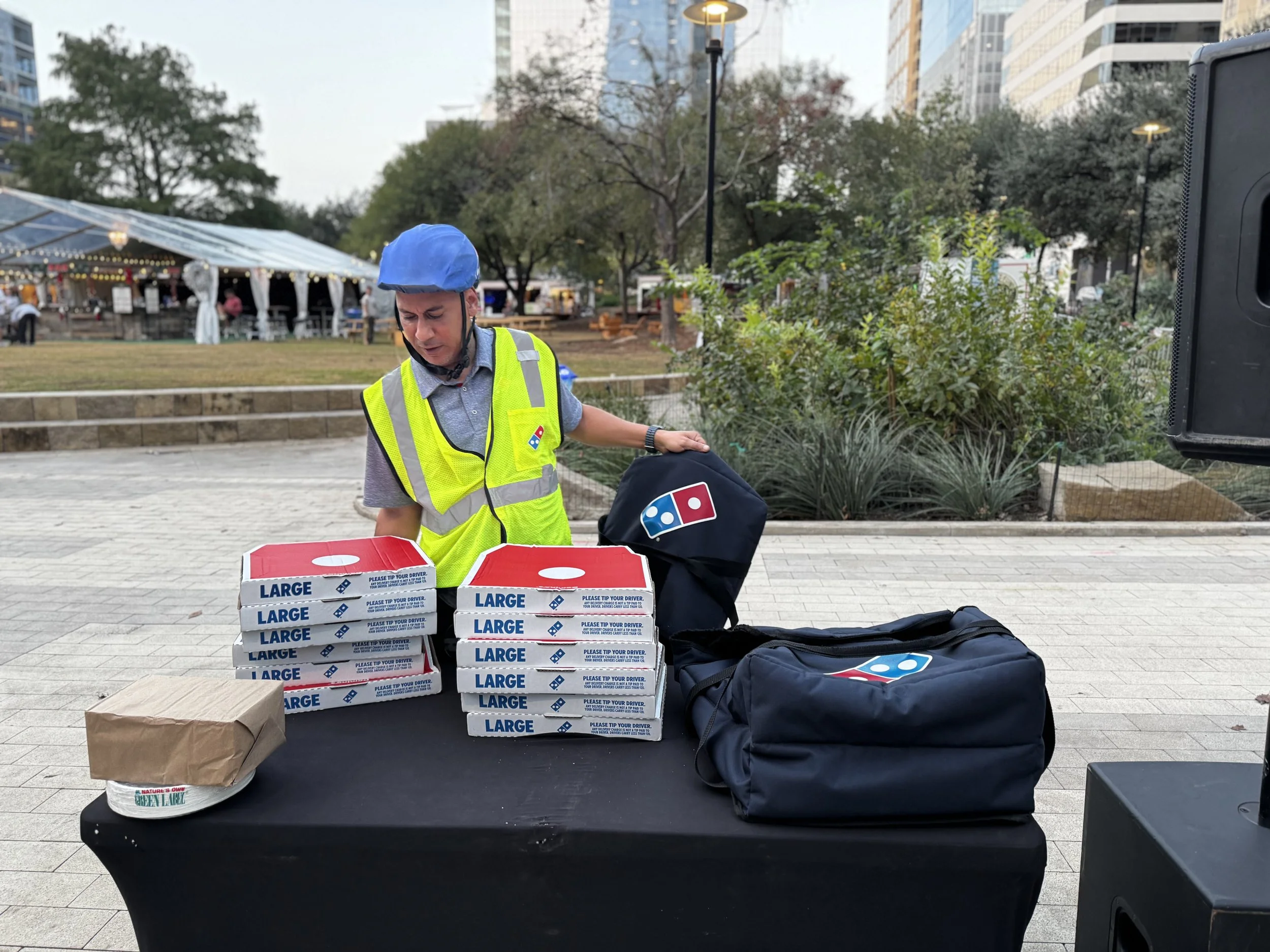 A person wearing a yellow safety vest and a blue helmet is preparing pizza boxes at an outdoor event. There are stacks of large pizza boxes labeled 'LARGE' on a black table, along with a small paper bag and a black bag with a Domino's logo. In the background, there are trees, a tent with string lights, and tall buildings.