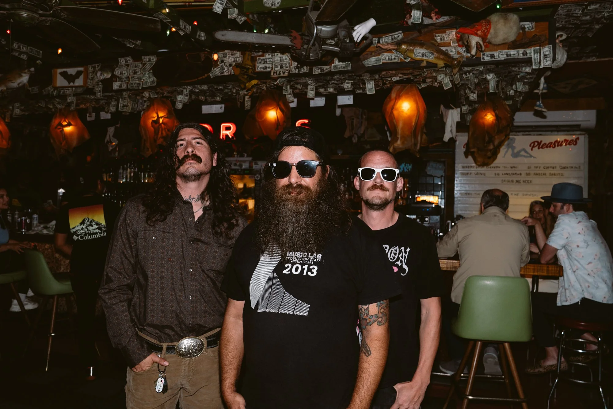 Three men standing together in a bar, with the ceiling decorated with hanging dollar bills and tied paper lanterns. The bar is lively, with other people socializing in the background.