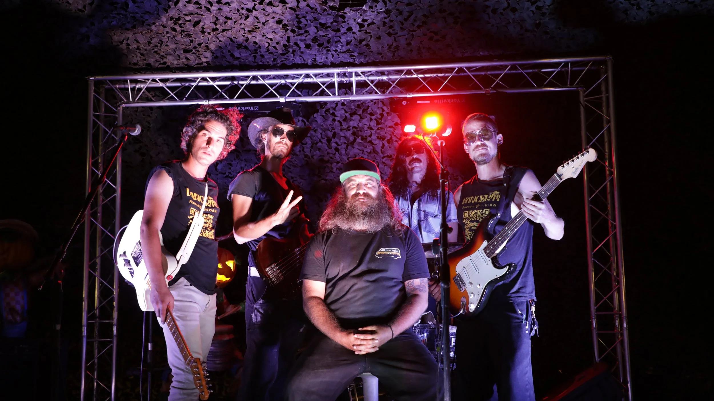 Group of five men posing with musical instruments on a stage at night, with a metal truss and colorful stage lights behind them.