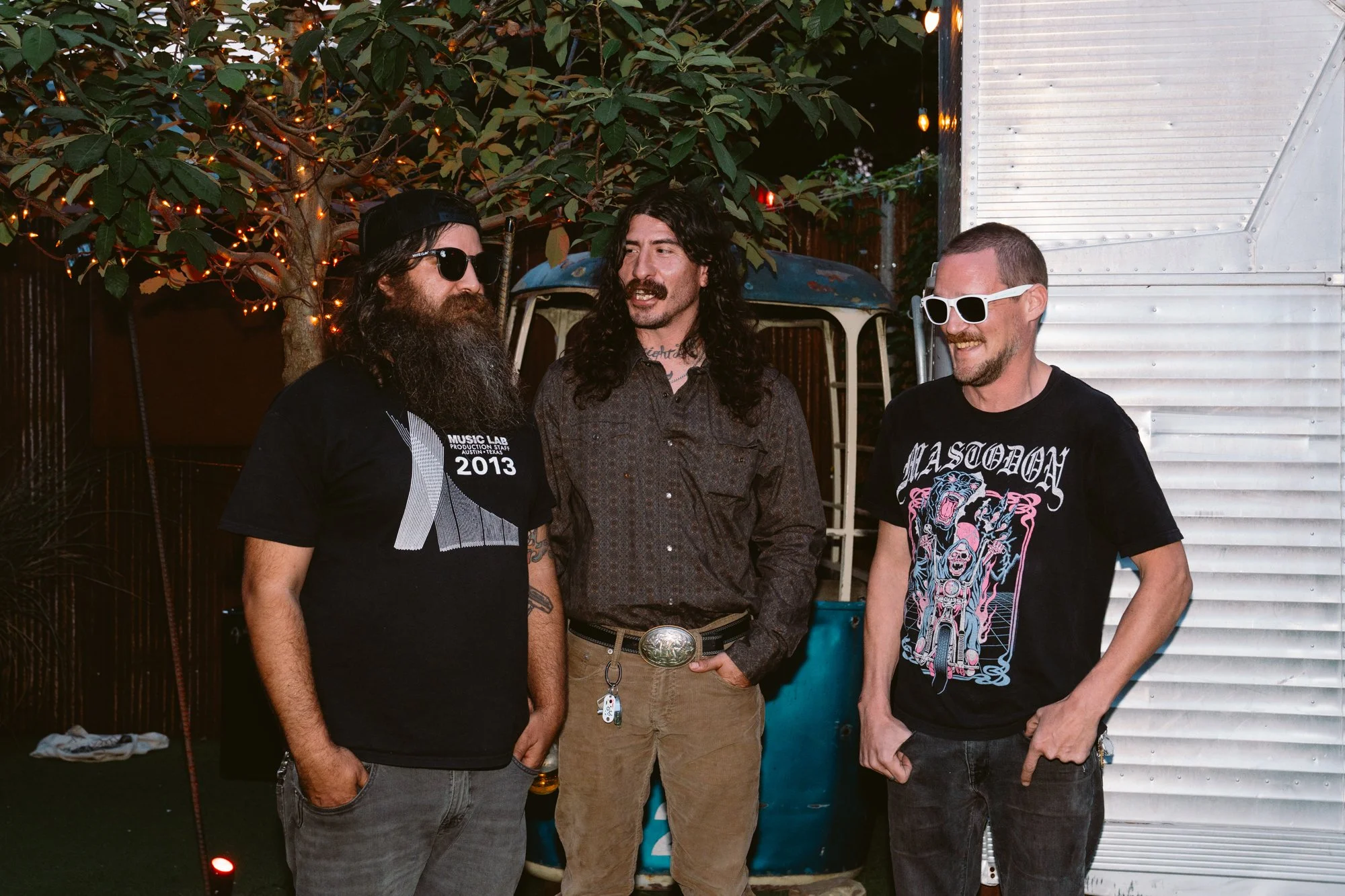 Three men standing outdoors near an old blue vehicle with a tree decorated with string lights in the background. The man on the left has long hair, a beard, sunglasses, and is wearing a black T-shirt. The man in the middle has long curly hair, a mustache, and is dressed in a dark patterned shirt. The man on the right has short hair, a beard, and is wearing white sunglasses and a graphic T-shirt.