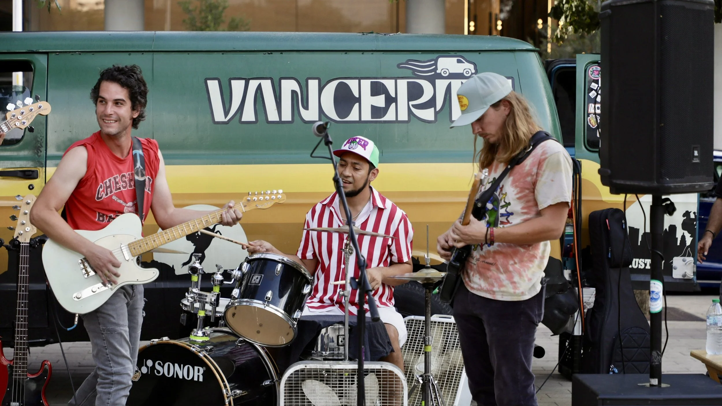 A group of young musicians performing outdoors with a green Vancer van in the background. The band includes a guitarist, a drummer, and a bassist, all casually dressed.
