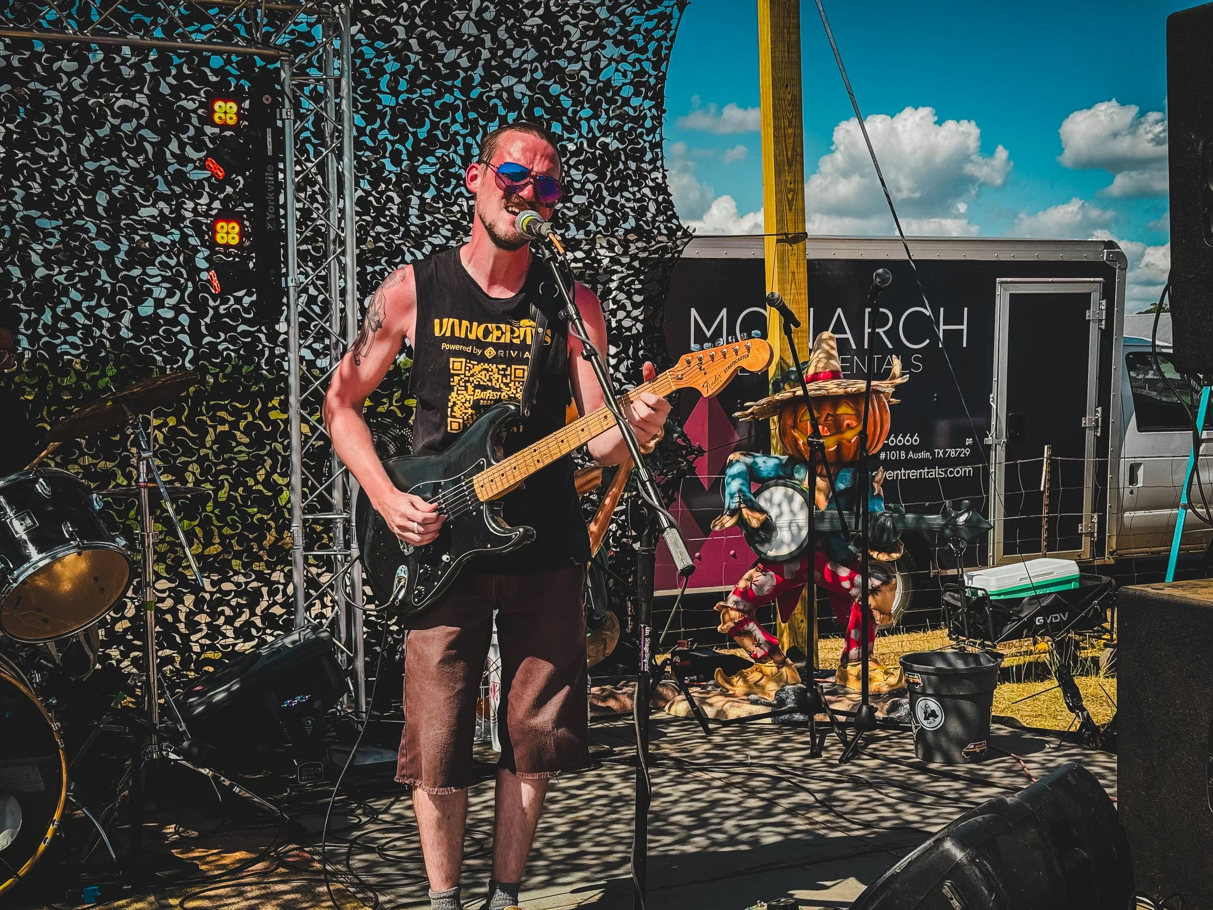 Male musician wearing sunglasses shirtless, playing an electric guitar and singing into a microphone on an outdoor stage during daytime, with a partly cloudy sky, decorated with a pumpkin sculpture in the background.
