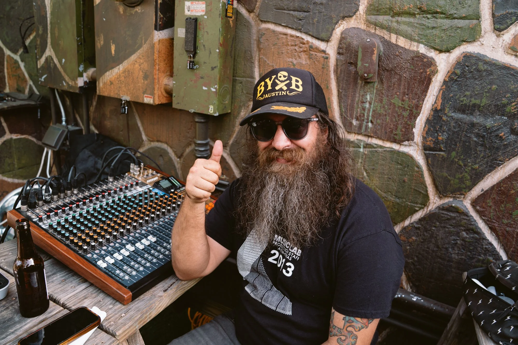 A man with a long beard and glasses wearing a black cap and T-shirt, giving a thumbs up while sitting at a table with a DJ mixer. The man is smiling and wearing dark sunglasses. The setting appears to be outdoors against a stone wall. There are beer bottles, a phone, and audio equipment on the table.