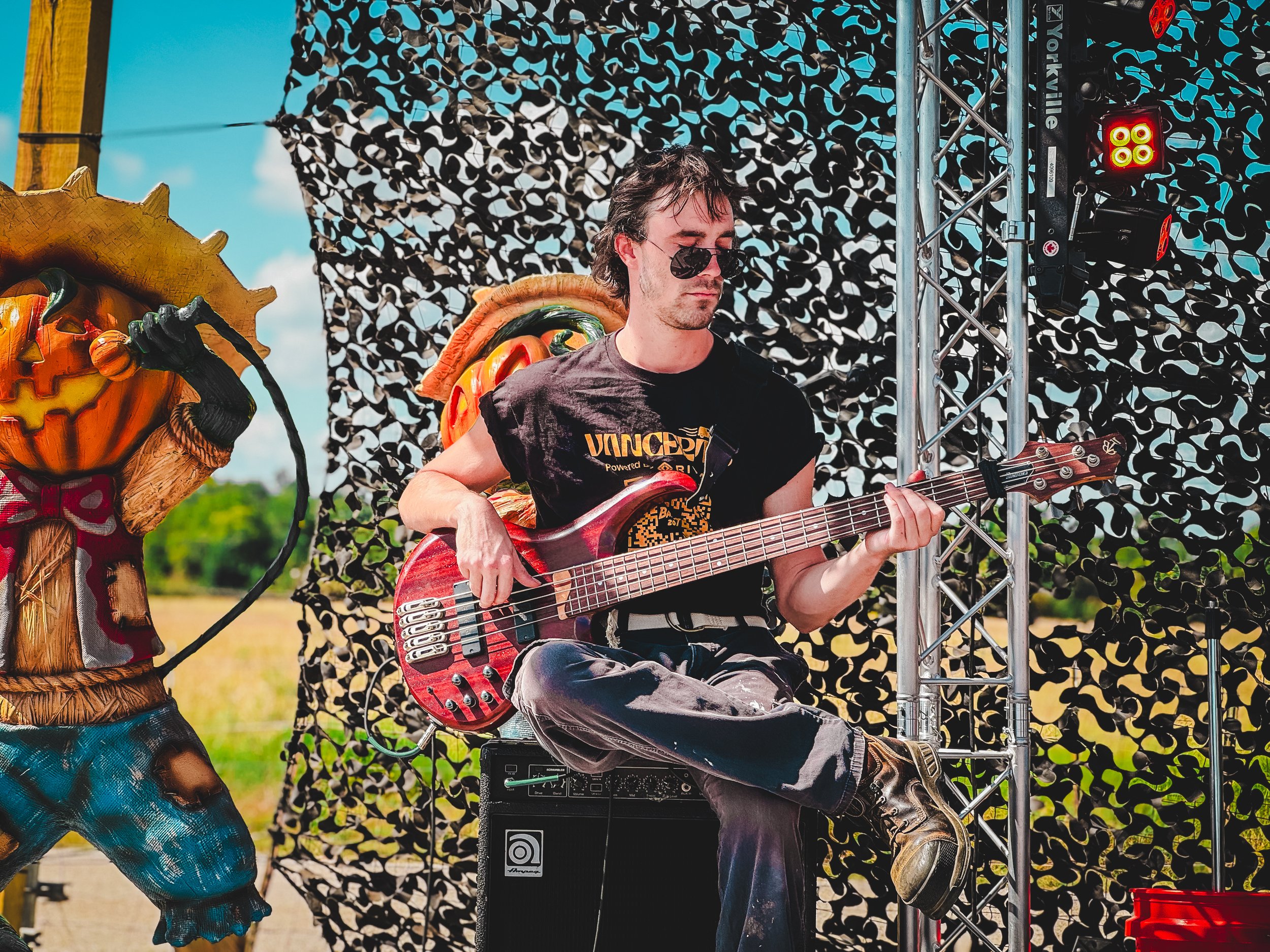 A man playing a red electric bass guitar on an outdoor stage with a decorative backdrop and a scarecrow figure nearby.