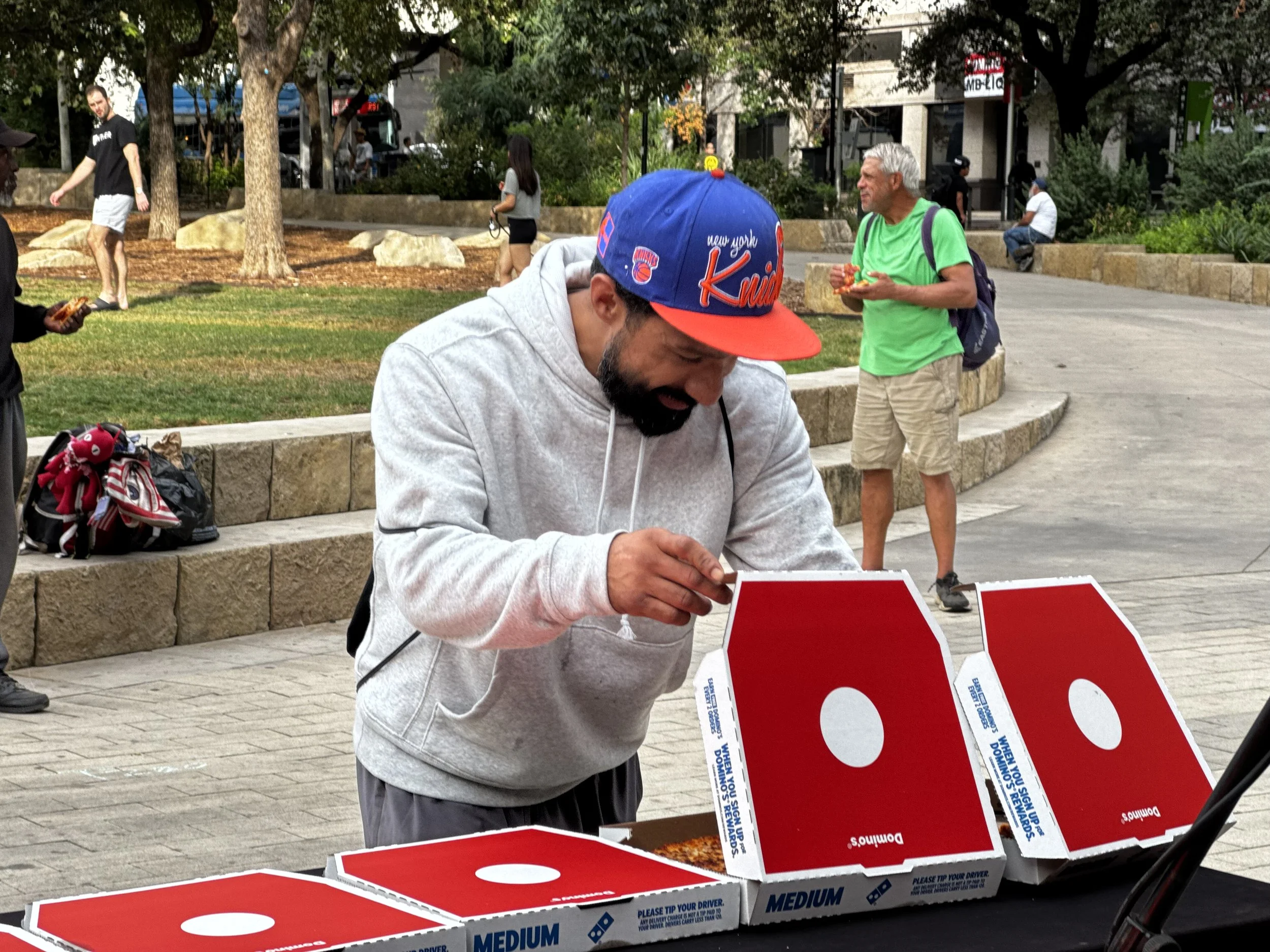 A man with a beard in a gray hoodie and a New York Knicks cap is leaning over a table of pizza boxes, inspecting a pizza inside one box. The table has multiple pizza boxes, some open, with red and white branding. In the background, people are walking in a park, some sitting on stone benches and others standing near trees.