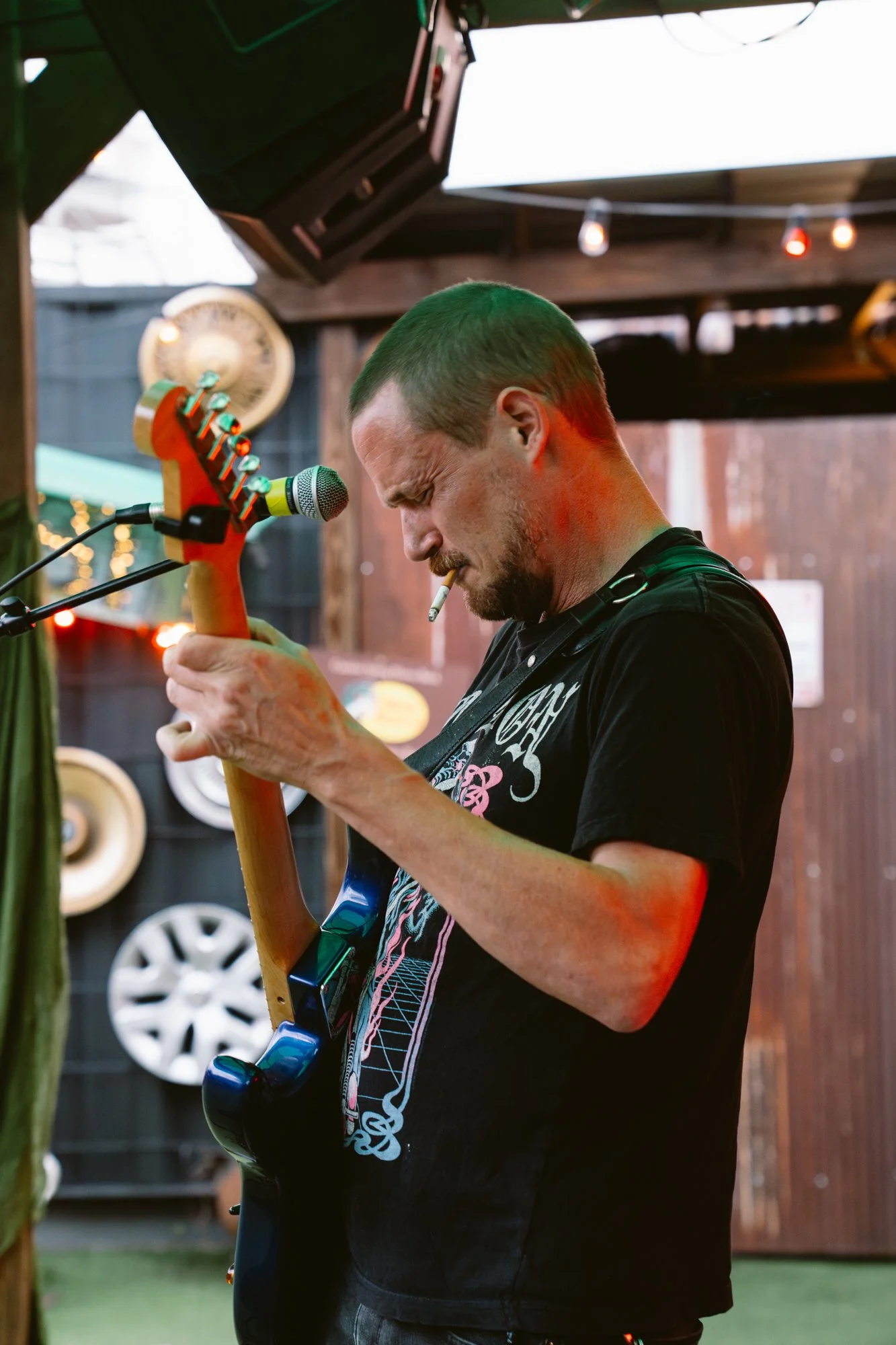 A man with a green mohawk hairstyle, cigarette in mouth, playing an electric guitar, standing in a rustic setting with string lights and car rims on the wall.