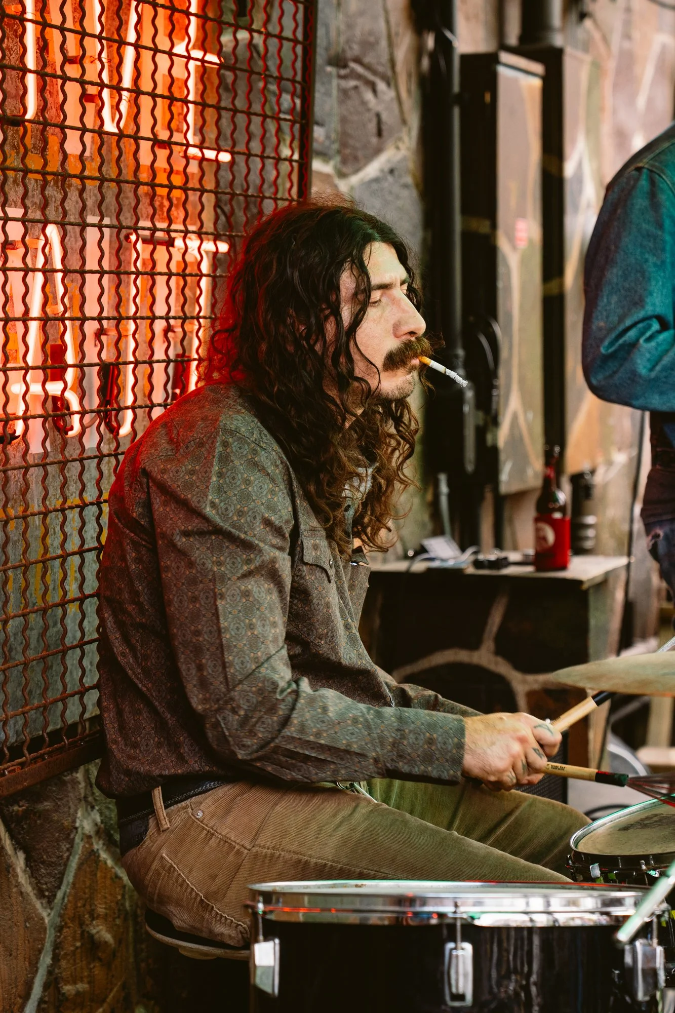 A man with long curly hair and a mustache, wearing a patterned shirt and brown corduroy pants, is sitting and playing drums while smoking a cigarette in a dimly lit, rustic setting with a wire mesh on the wall behind him.