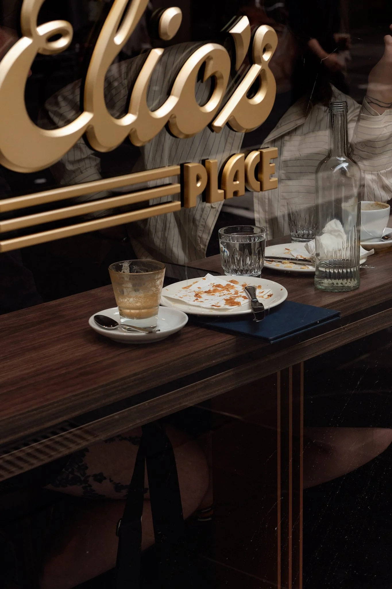 View through a window showing a vintage neon sign that spells 'Good' with reflection of people and dining table inside, including empty plates, a spoon, a glass bottle, and partially visible person.