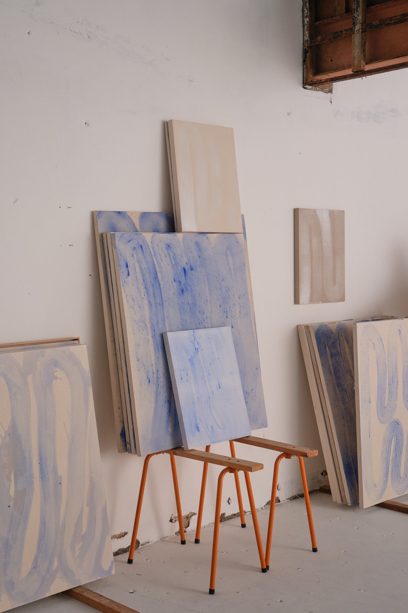 A room with multiple canvases featuring abstract blue and white swirl designs, propped against the wall and on orange sawhorses, with visible construction or renovation details.