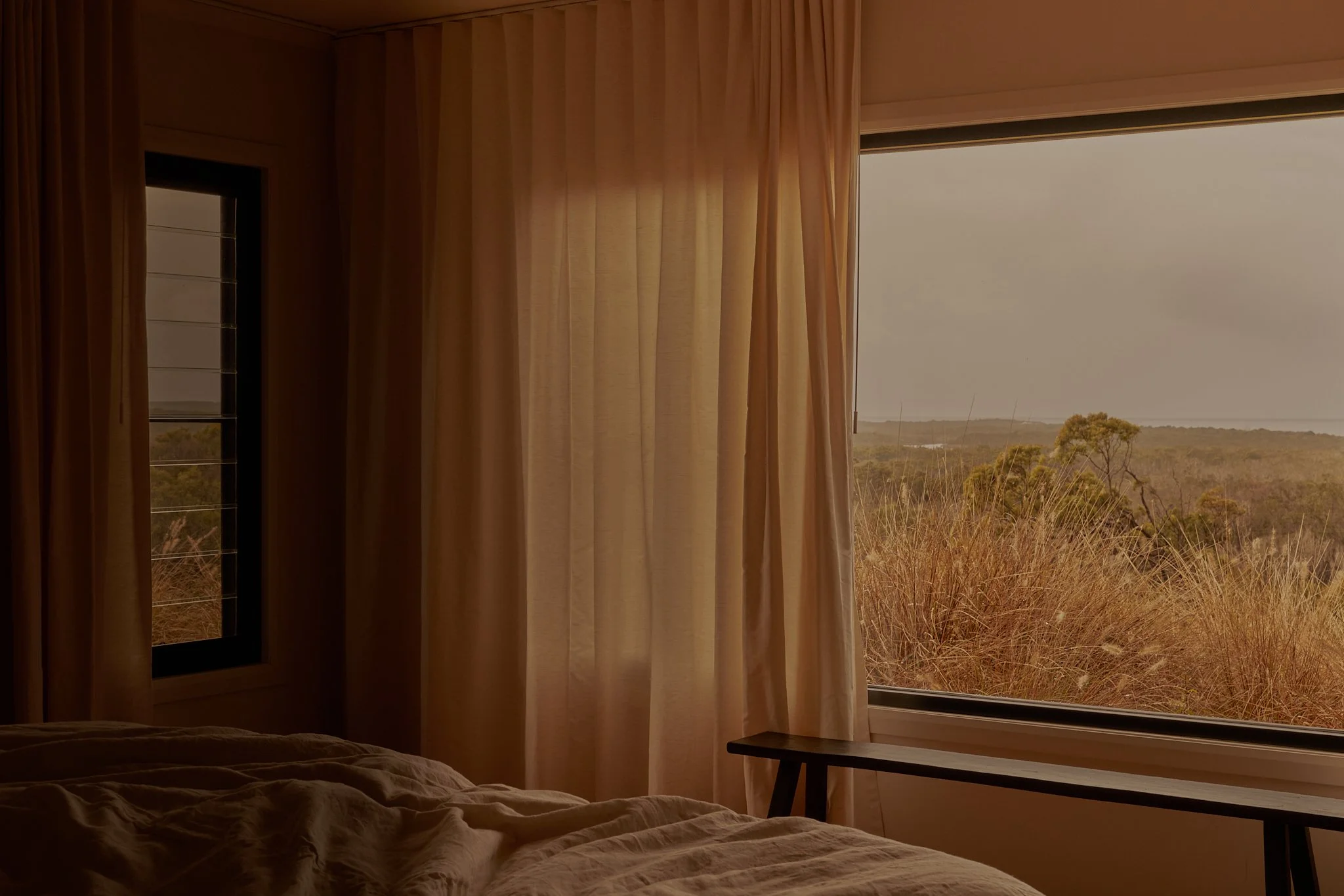A bedroom with beige curtains partially open, revealing a view of tall grasses and trees outside on a cloudy day. There is a bed with rumpled bedding in the foreground and a small table or bench underneath the window.