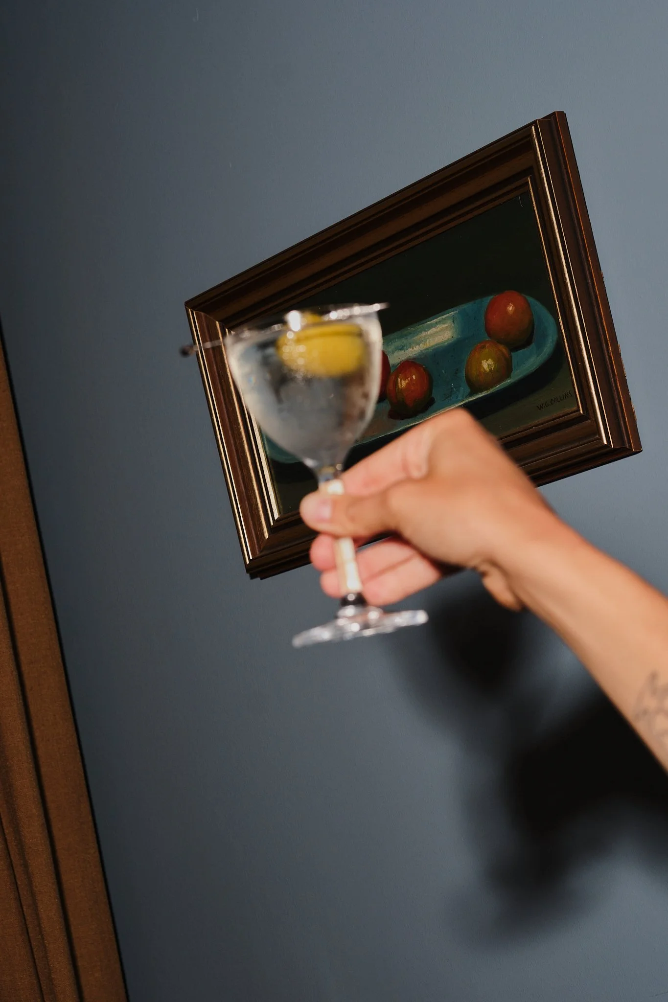 Close-up of a hand holding a cocktail glass with lemon, in front of a framed still life painting of apples on a blue oval plate.
