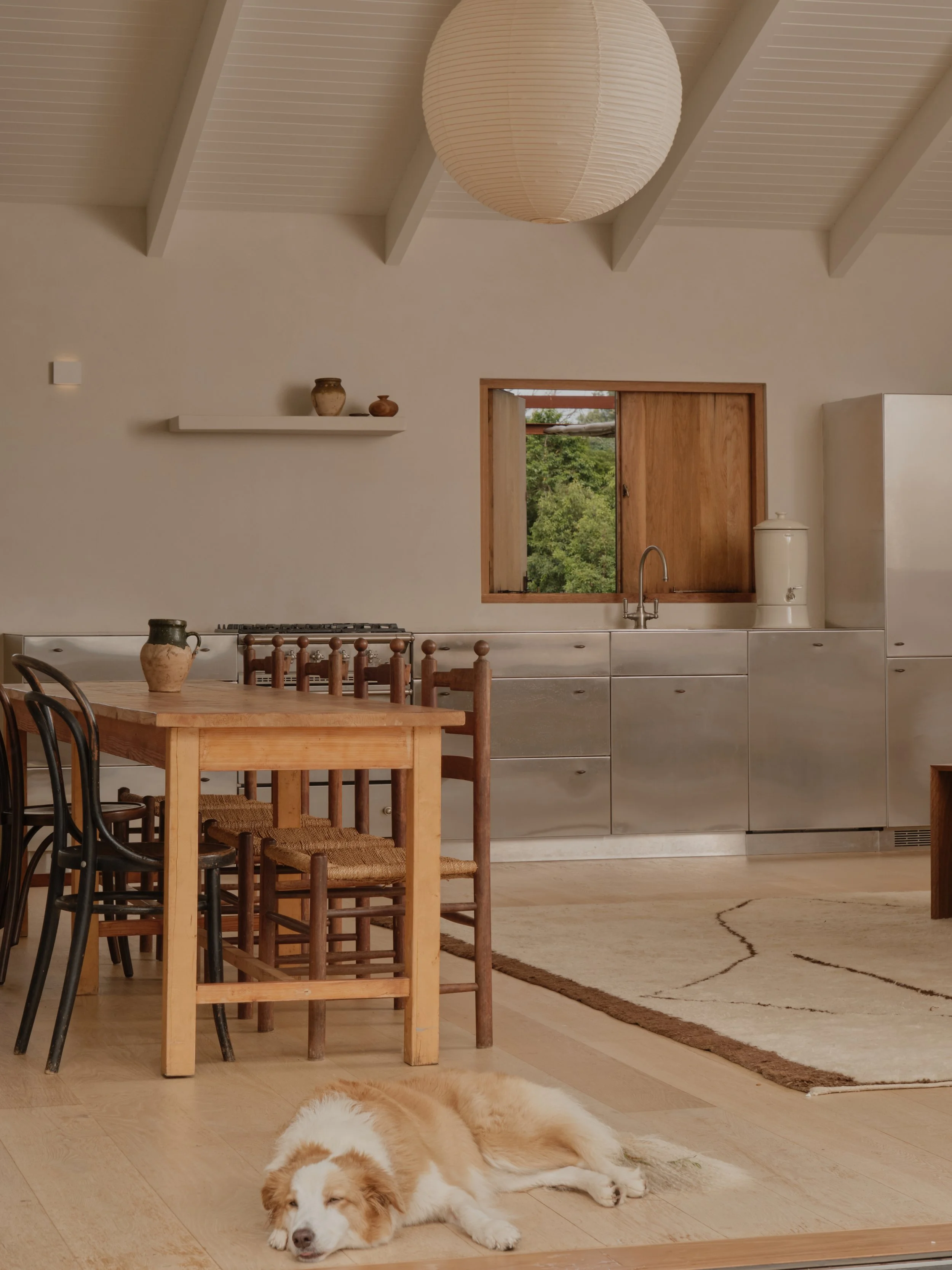 A cozy kitchen with a wooden dining table and black chairs, beige cabinets, a window showing greenery outside, a fluffy white and brown dog lying on the floor, a paper lantern hanging from the ceiling, and decorative vases on a shelf.