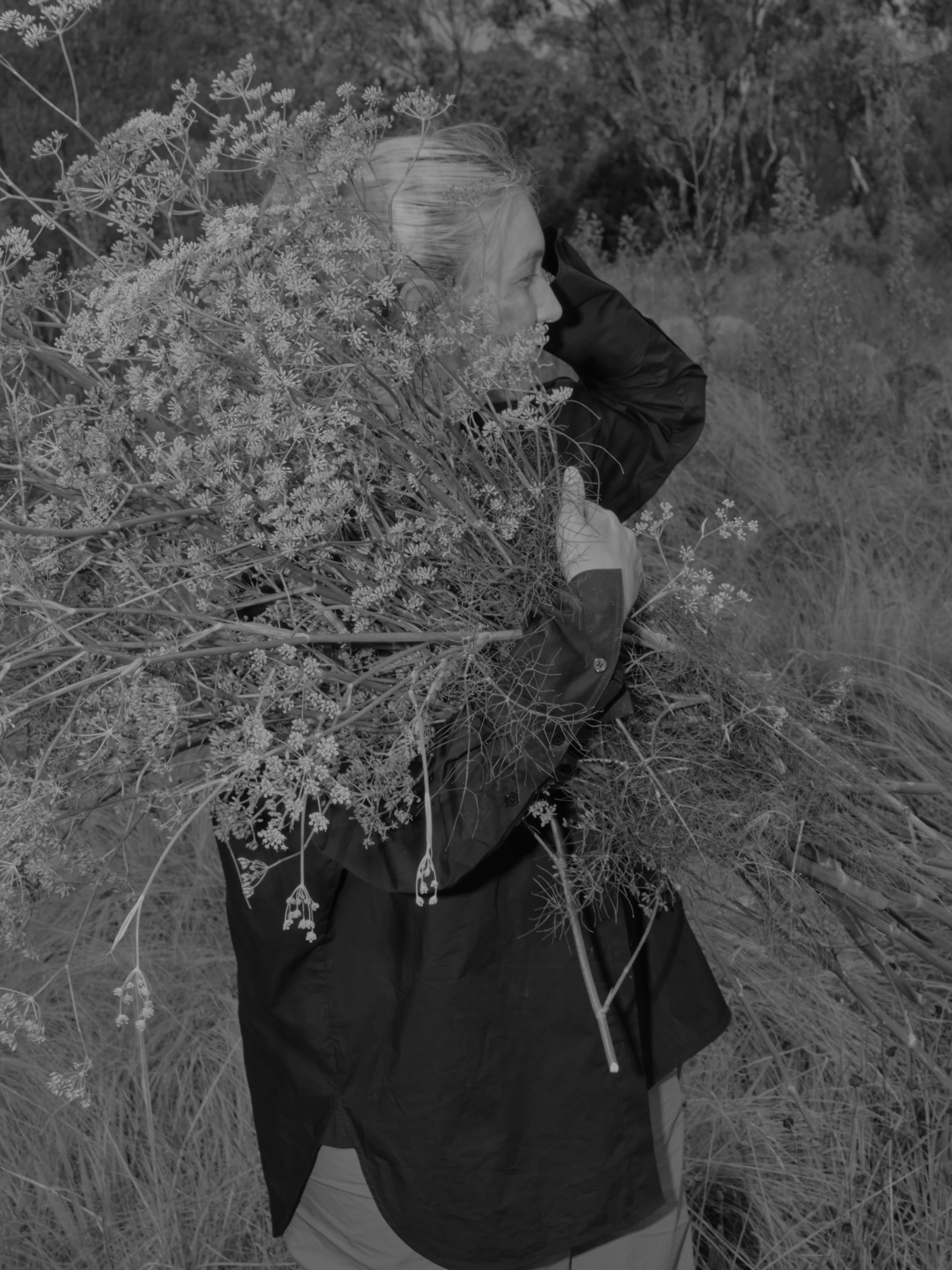 Person with blonde hair in a black jacket and light-colored pants, sitting in tall grass and near bushes with flowers, hands on head in an outdoor setting.