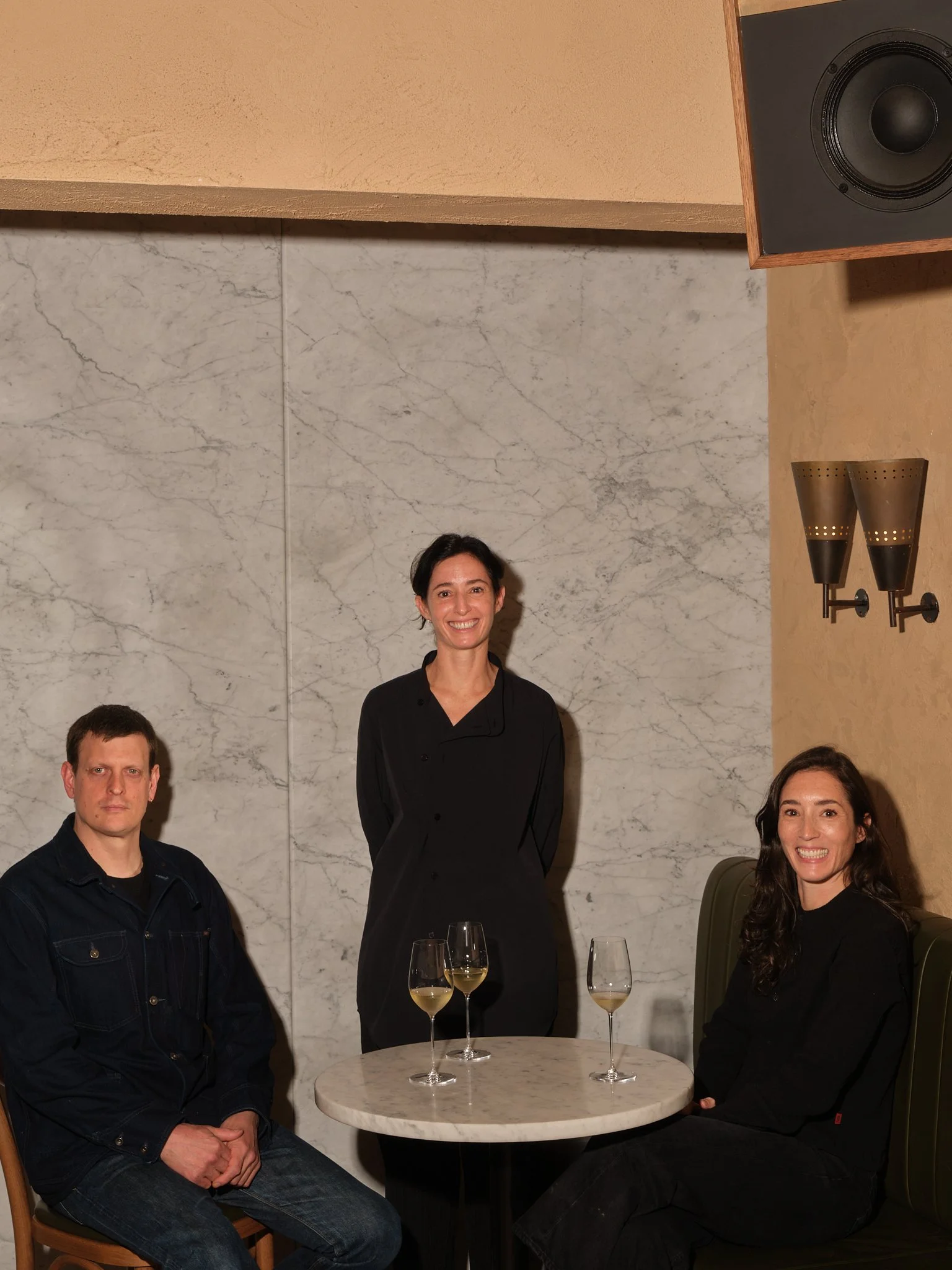 Four people, three women and one man, sitting and standing around a small round table with three glasses of white wine, in a restaurant with a marble wall background and a speaker on the wall.