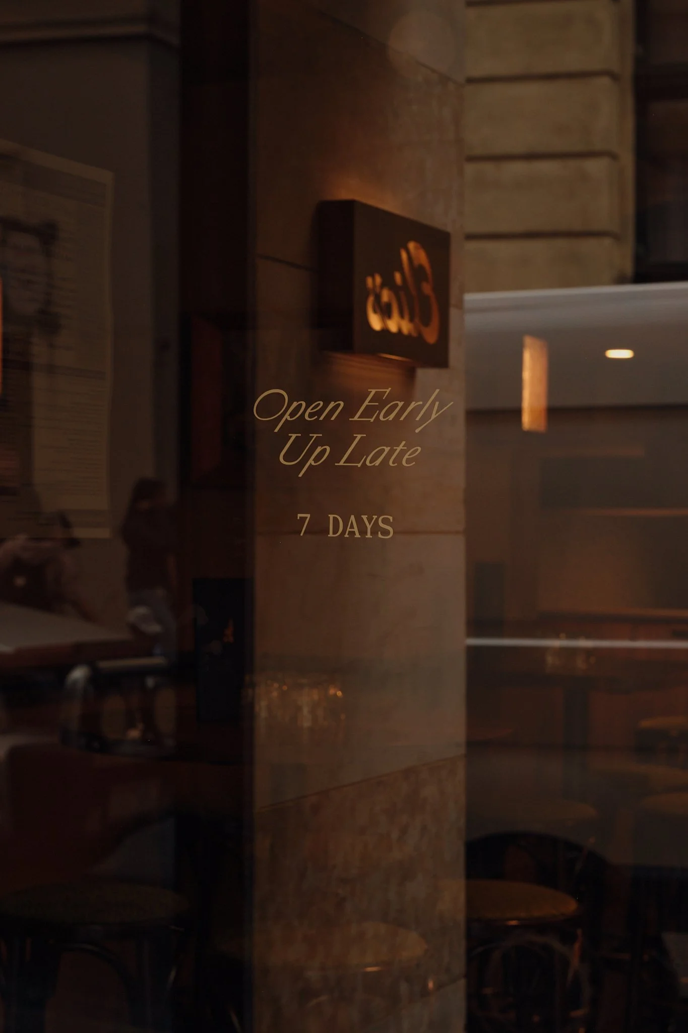 A restaurant window with a sign that reads 'Café' lit from behind, and an overlay of text that says 'Open Early Up Late 7 Days.'