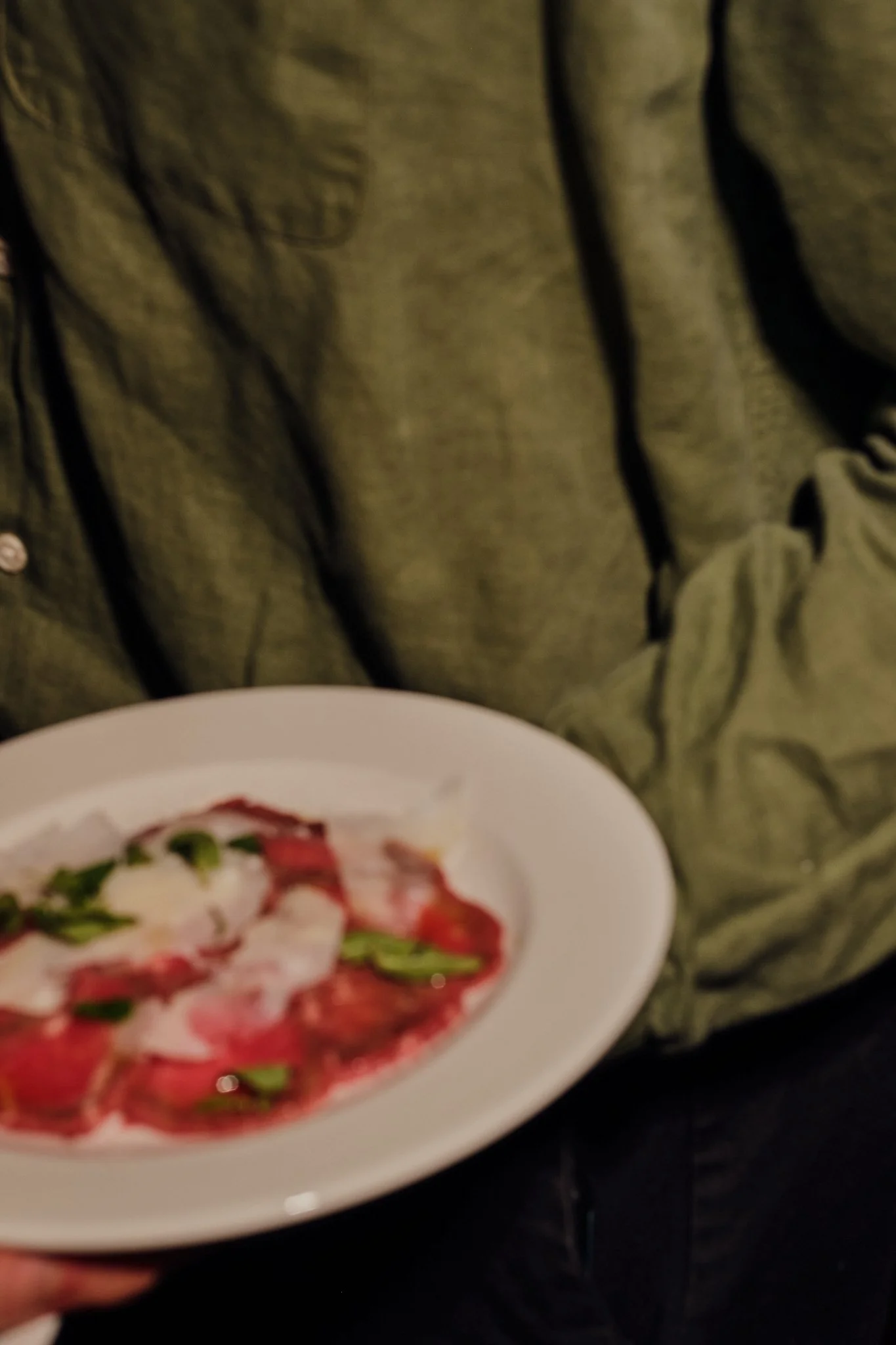 A plate of salad with sliced tomatoes, fresh greens, and mozzarella cheese, being held in front of a person wearing a green jacket.