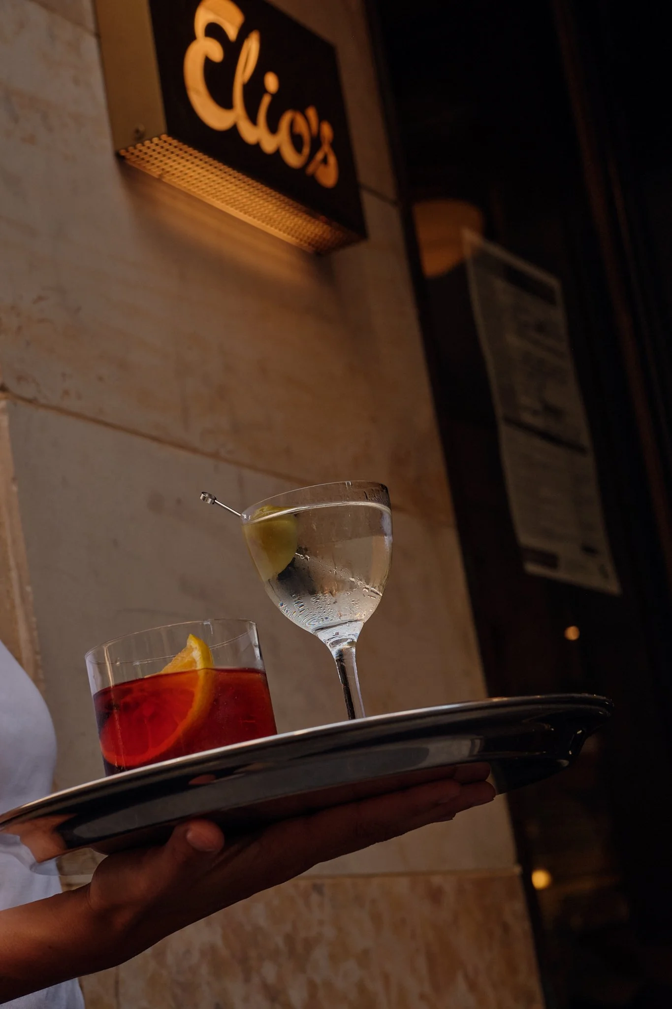 A tray with three cocktails, one with an orange slice and the other with a green olive, possibly lemon, in a bar setting with a sign for 'Elirio's' in the background.