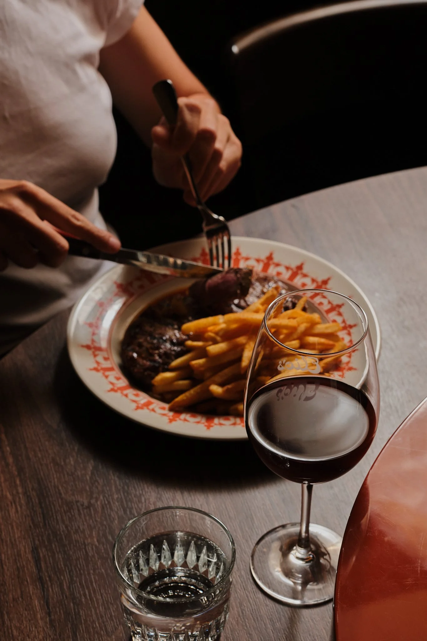 A person with a white shirt is cutting a steak on a plate of French fries, with a glass of red wine and a glass of water on the table.