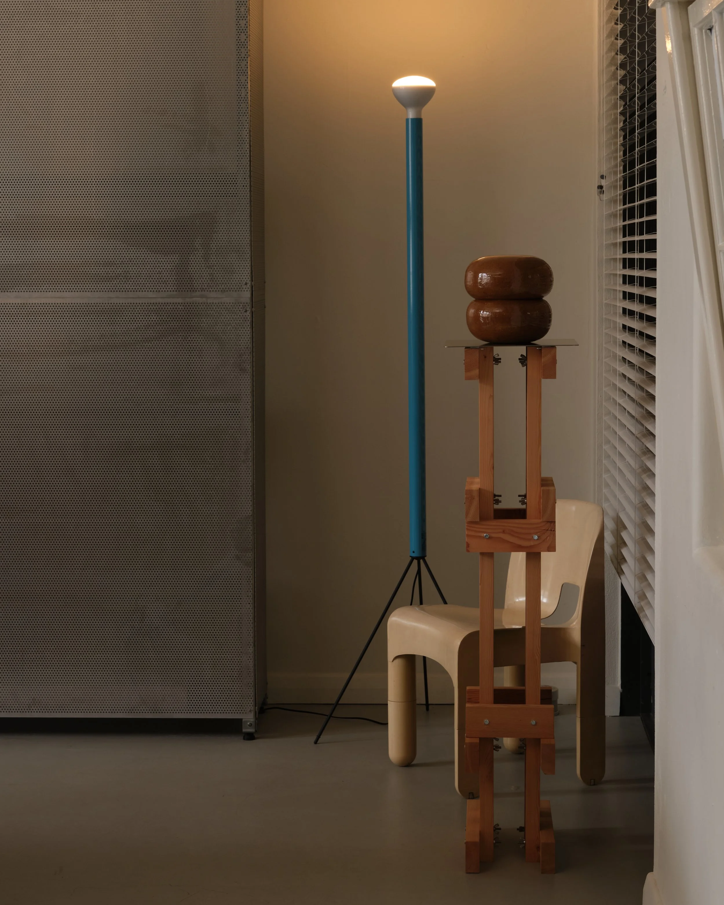 A scene with a floor lamp emitting warm light, next to a wooden table with a wooden sculpture or object on it, chair, and a modern metallic cabinet on the left side. There are also white window shutters on the right side.