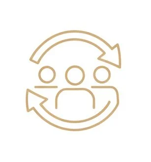 A woman with long wavy hair in a blue blazer and tan top, smiling and leaning against a white wall in an office or classroom with tables, chairs, and windows in the background.