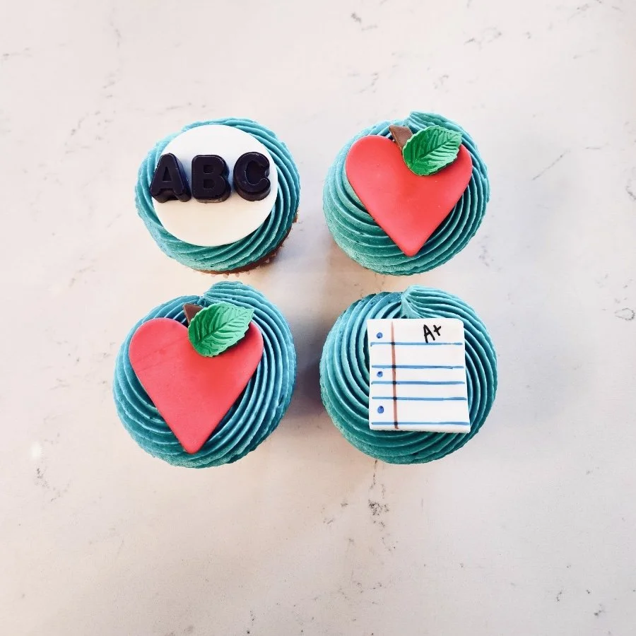 Four cupcakes with teal frosting, decorated with themes of school: "ABC" letters, a red apple with a leaf, and a page marked "A+."