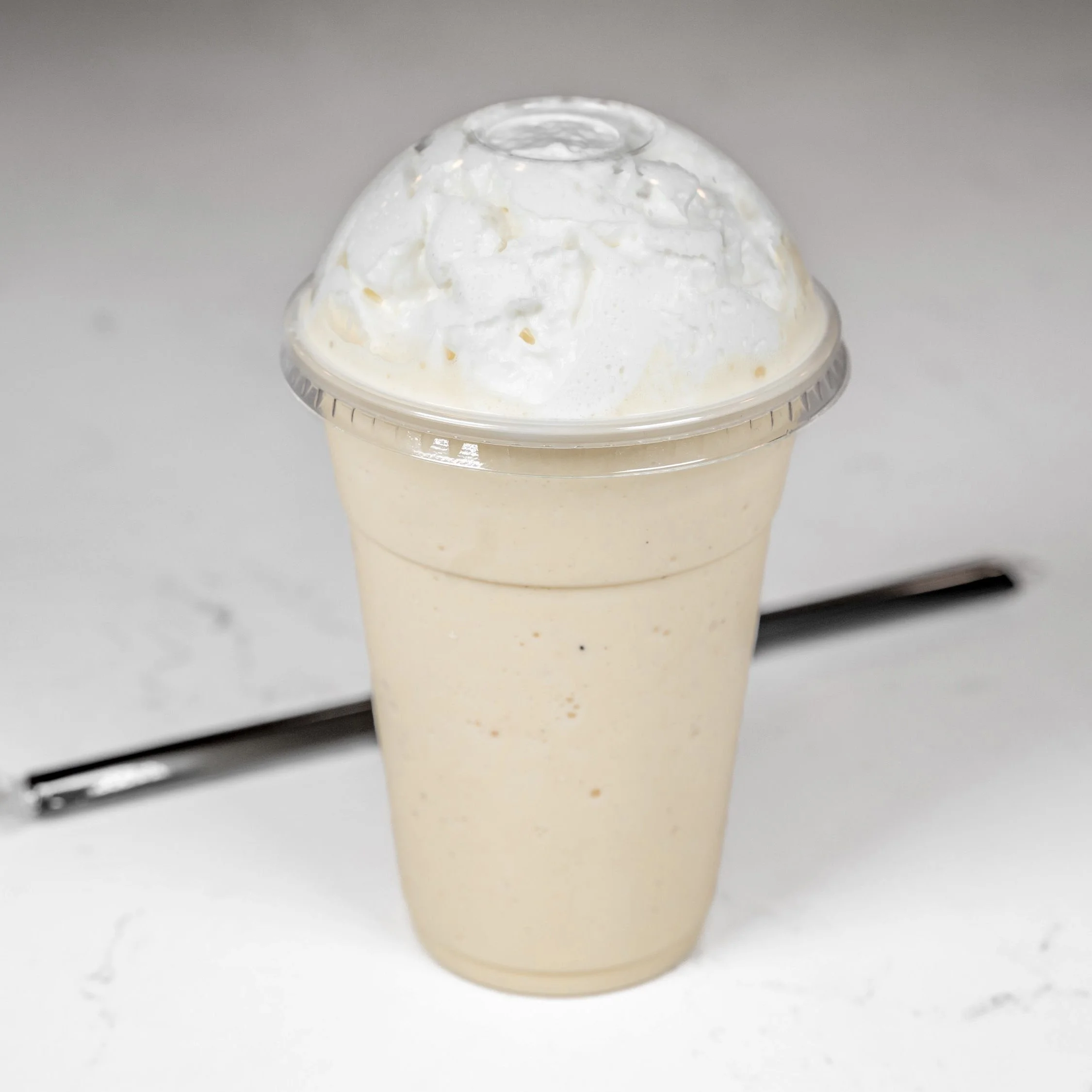 Vanilla milkshake in a plastic cup with whipped cream on top, placed on a white surface with a straw beside it.
