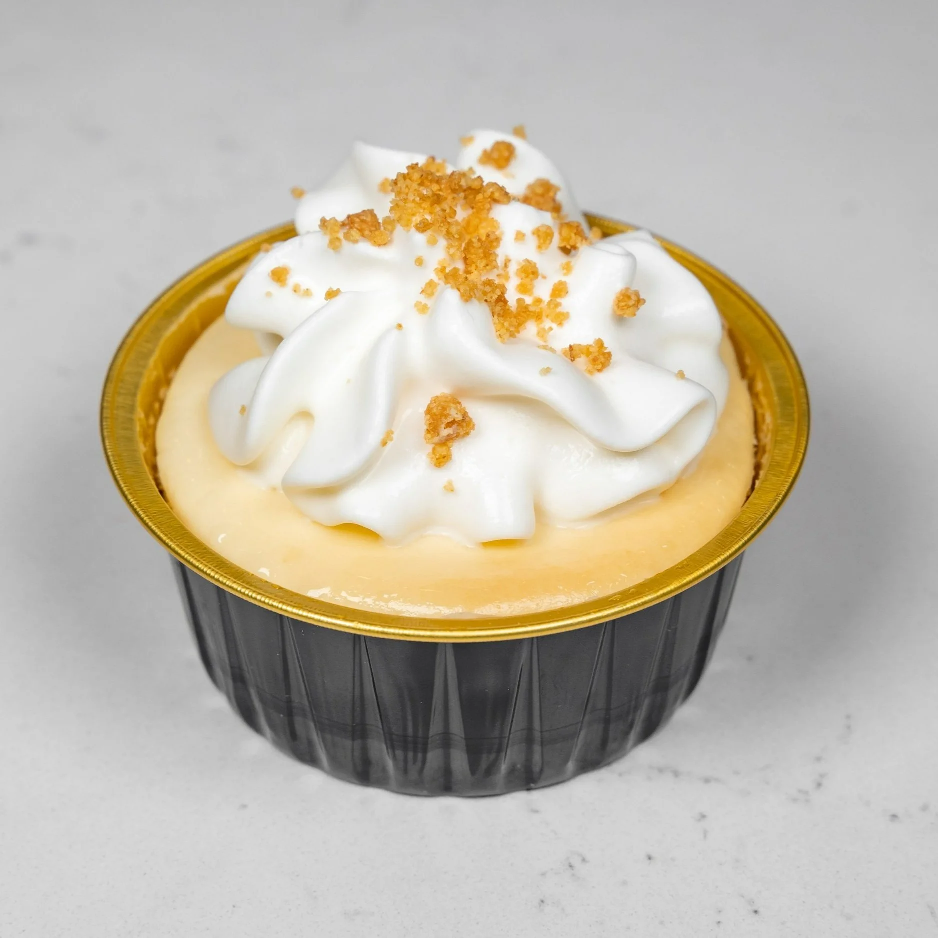 A creamy dessert in a black and gold cup, topped with whipped cream and sprinkled with crumbs, on a white surface.