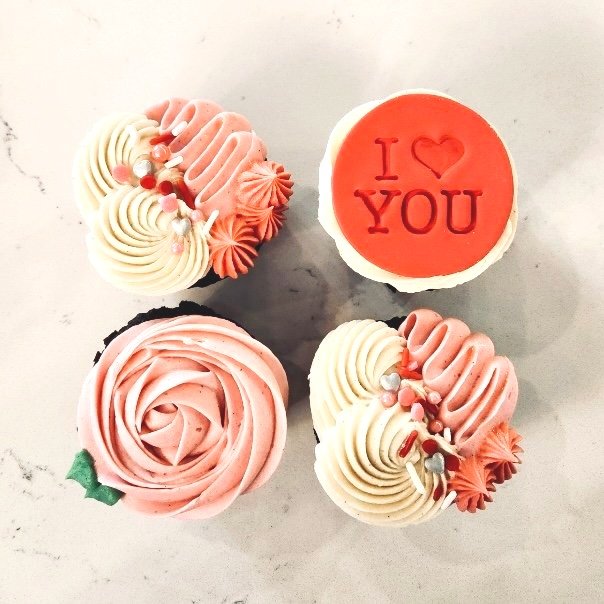 Four decorated cupcakes with pink and white frosting, one cupcake has a red topper with 'I ❤️ YOU' written on it.