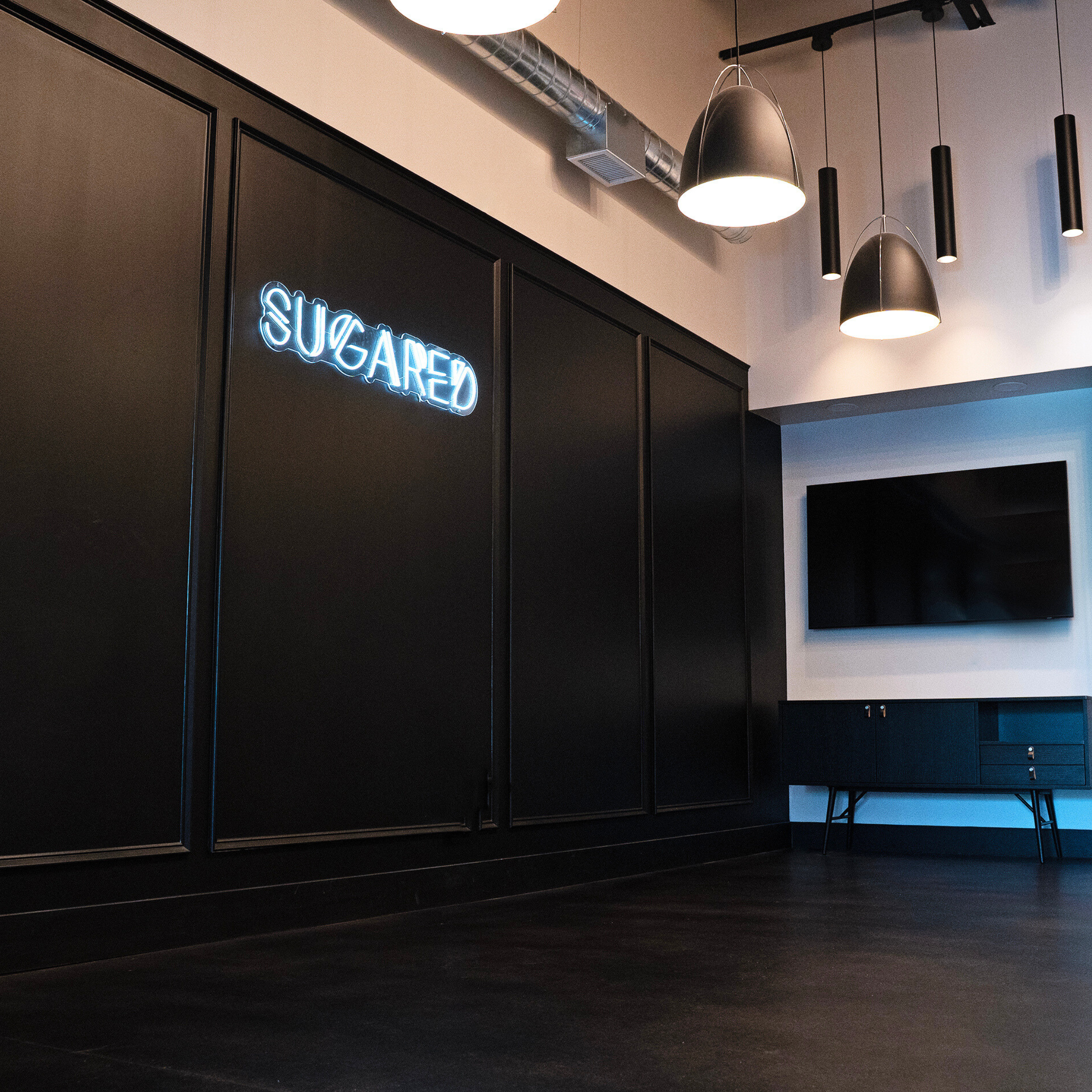 Modern interior with black wall panels, neon sign "Sugared," ceiling lights, and wall-mounted TV above a cabinet.