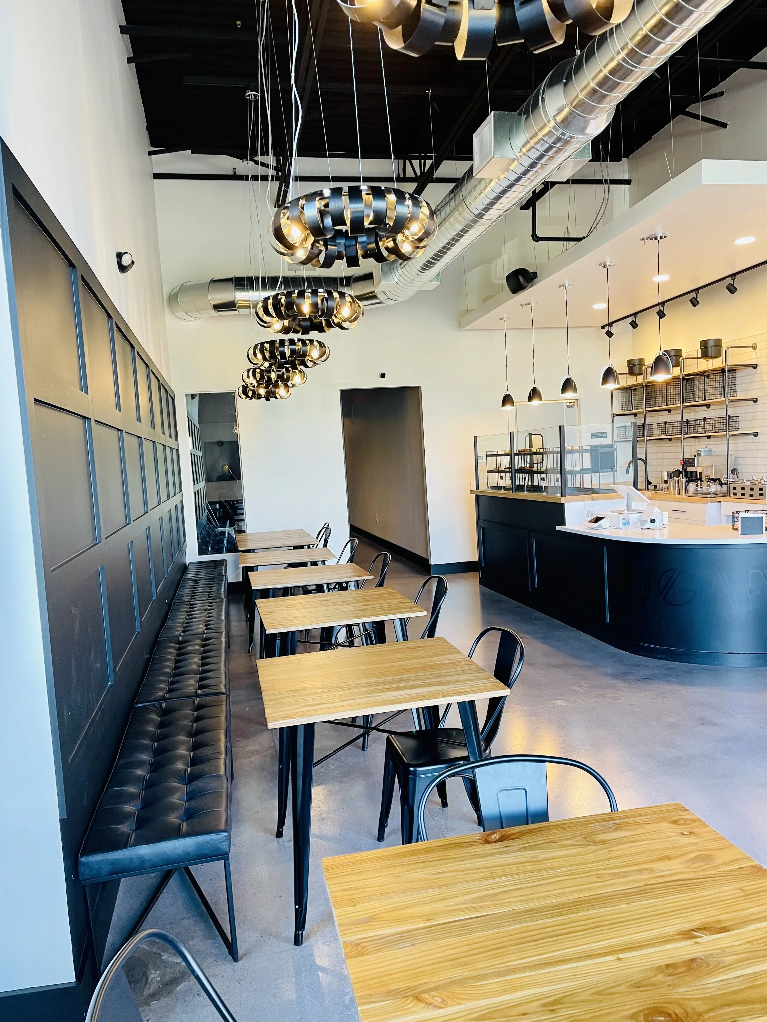 Modern dessert lounge interior with wooden tables, metal chairs, black tufted bench seating, and industrial-style pendant lights. The setting features a sleek counter area with shelves and minimalist design elements.