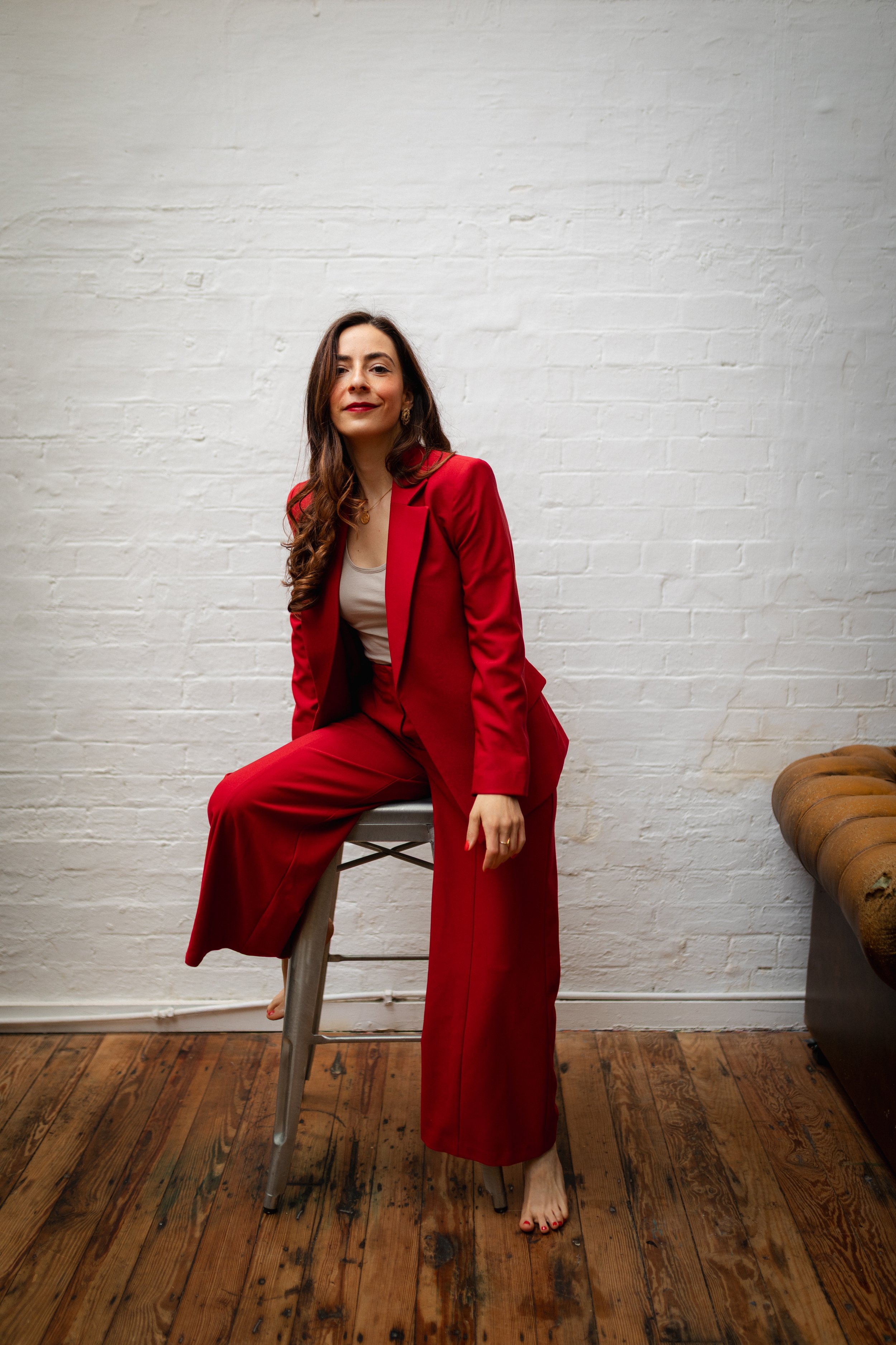 A woman in a red suit sitting on a chair in front of a white brick wall, with wooden flooring and a brown leather ottoman nearby.