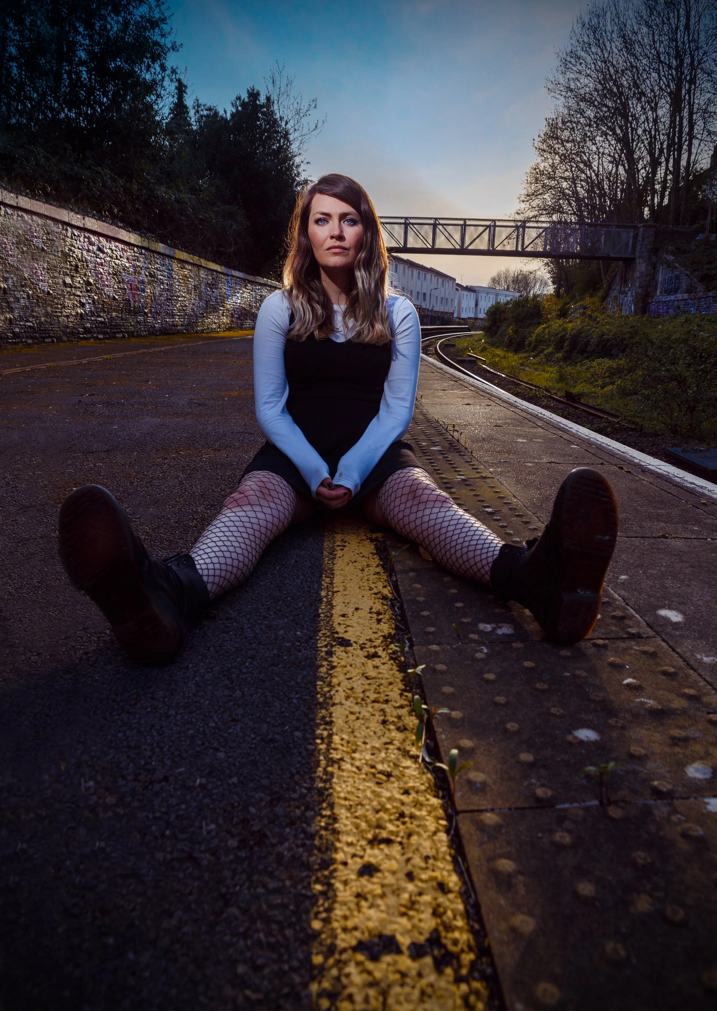 Shauna Tohill from the band Rews, poses sitting on the ground of a railway platform. A yellow painted line runs between her legs from the foregound into the distance. The sun is setting behind her in the blue sky.