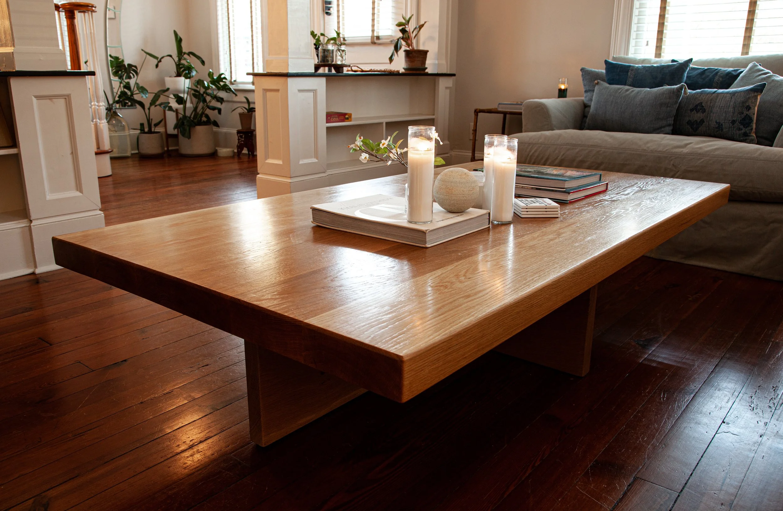 a large warmly finished white oak coffee table sits firmly in a cozy living with lush natural plants surrounding the background and plush blue pillows on the adjacent sofa