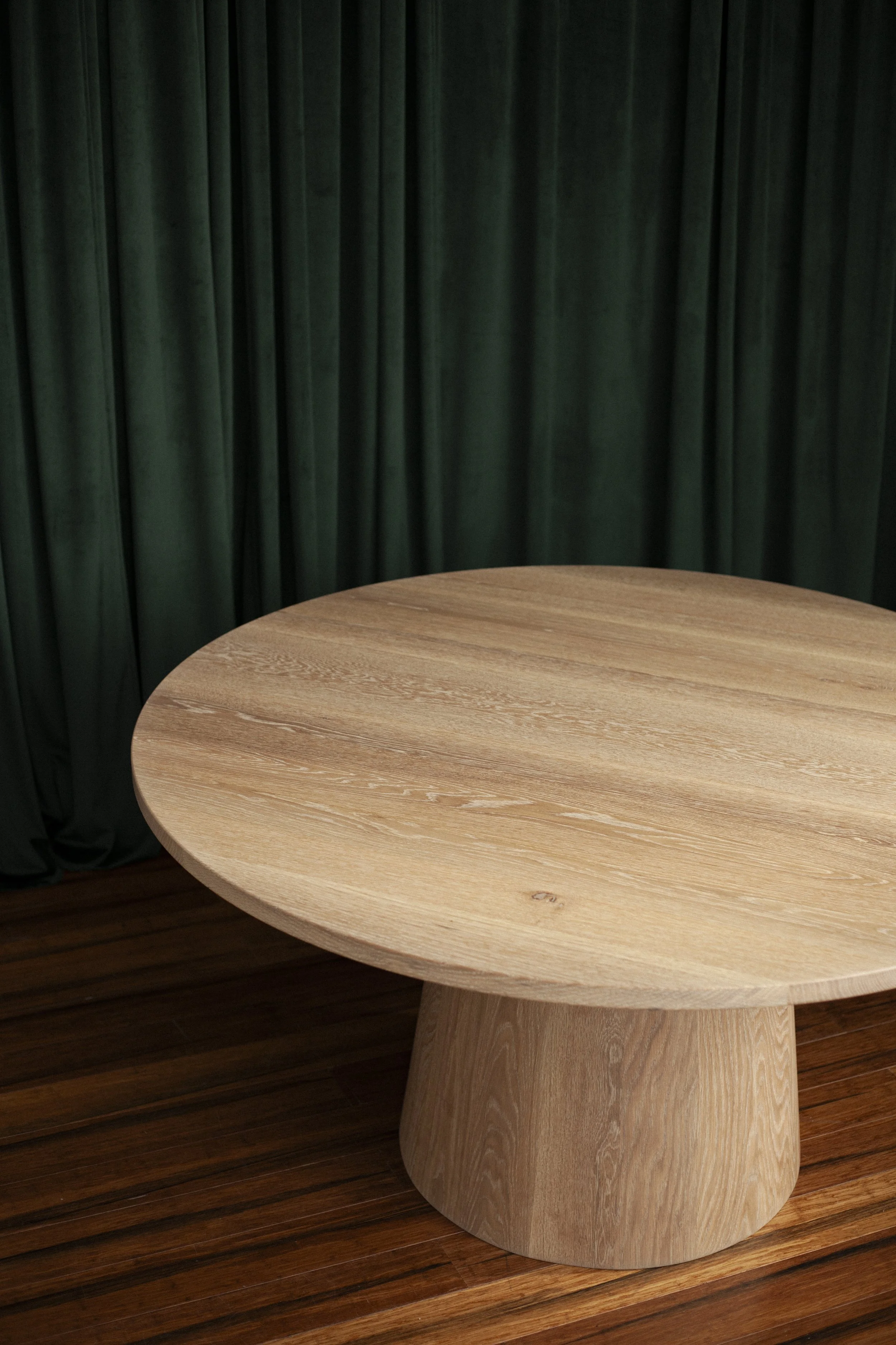 circular white oak pedestal dining table with a conical base sitting on rich hardwoods and dark green, velvety curtains as the background in a naturally lit studio space