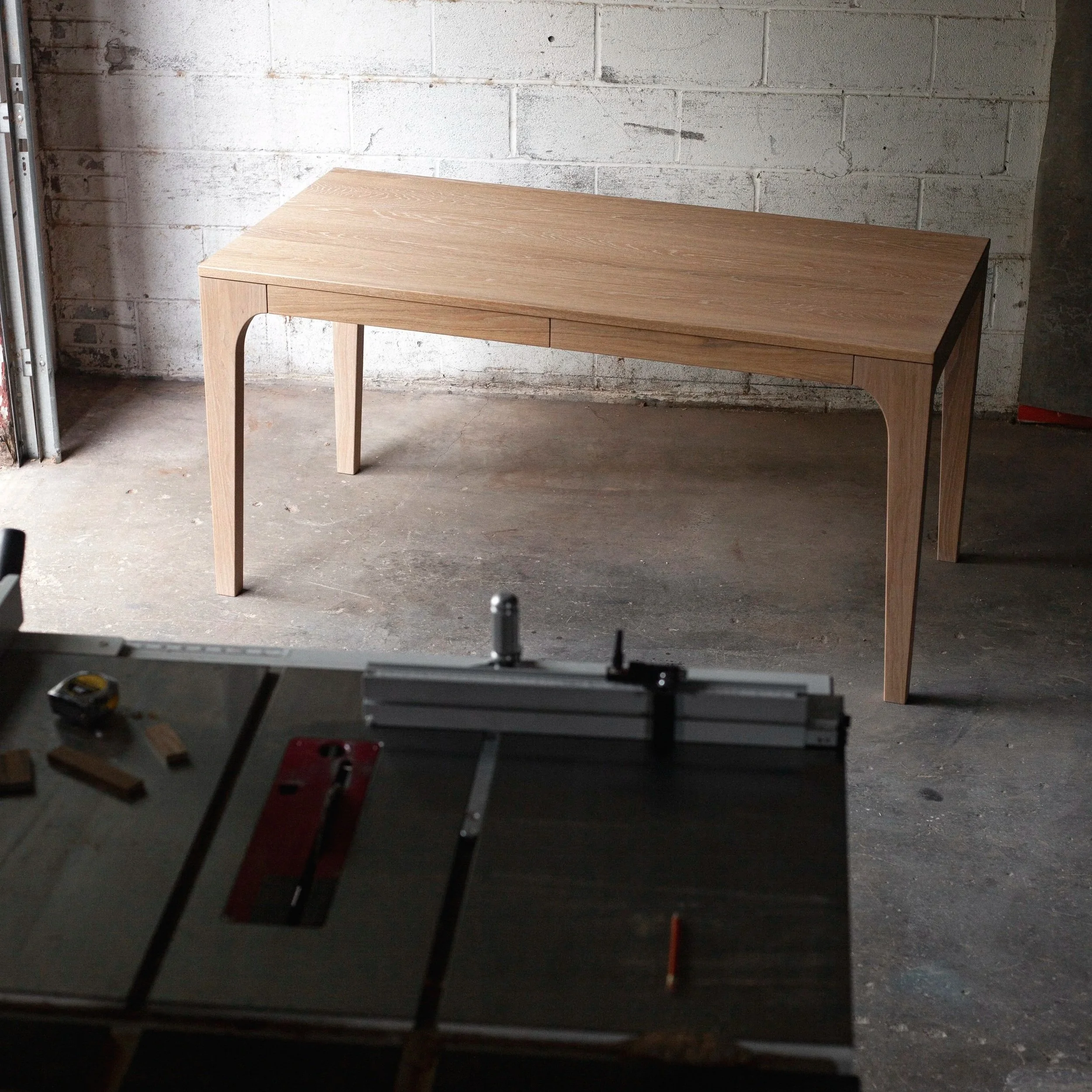modern white oak desk with two hidden drawers with a natural, warm color finish sitting on workshop floor with a tablesaw and measuring tools in foreground