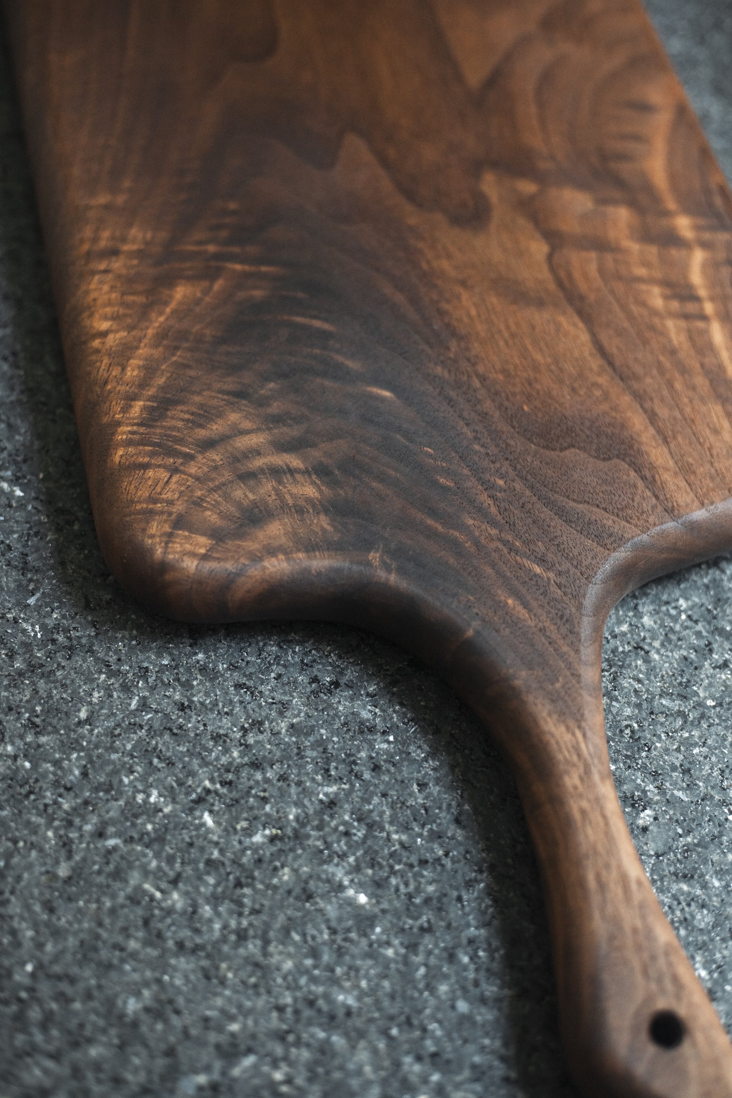 highly figured walnut charcuterie serving board with a sword shaped handle sitting on a dark granite countertop