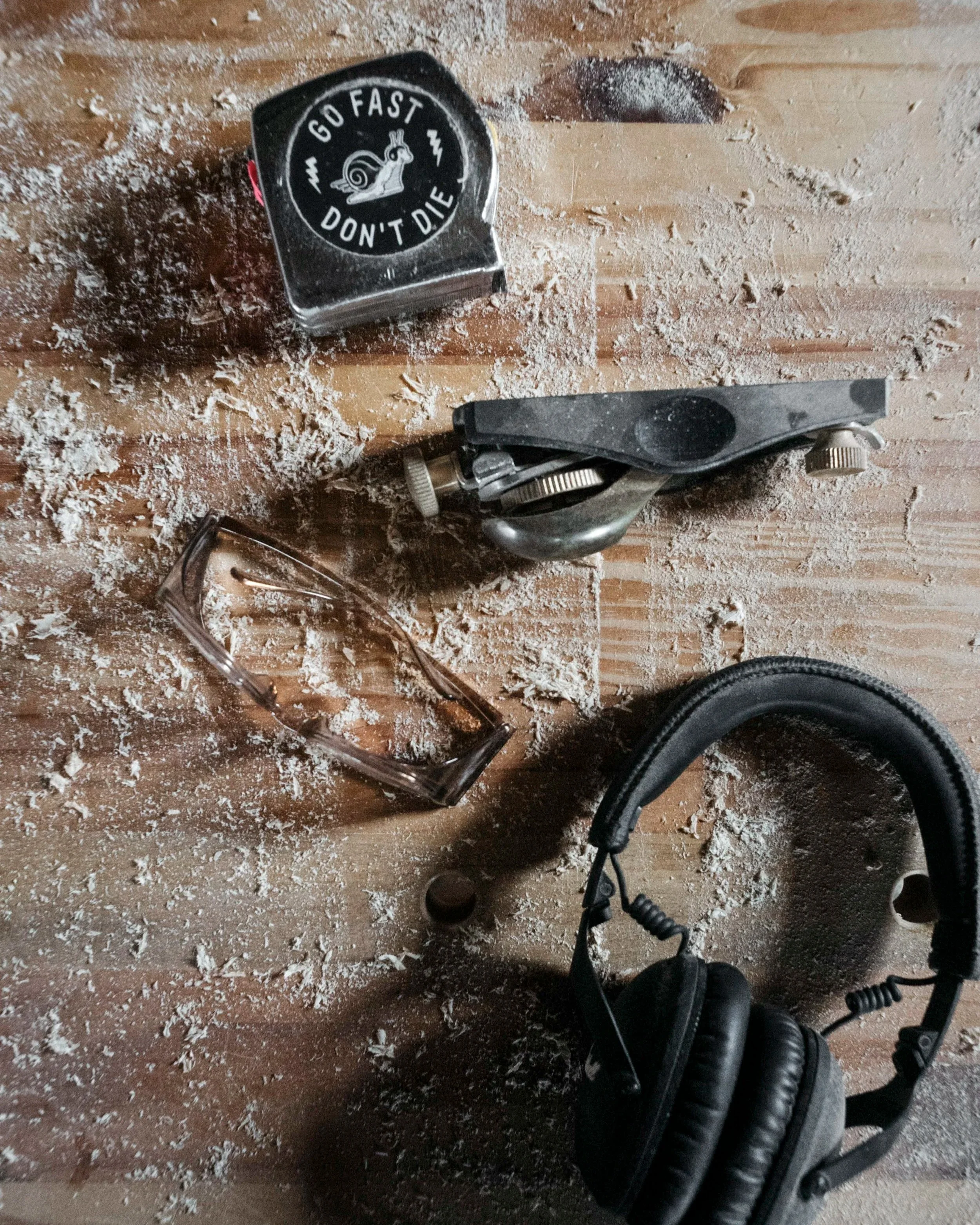 a block plane, tape measure, safety glasses, and headphones sit on a dusty workbench surface with cool natural light
